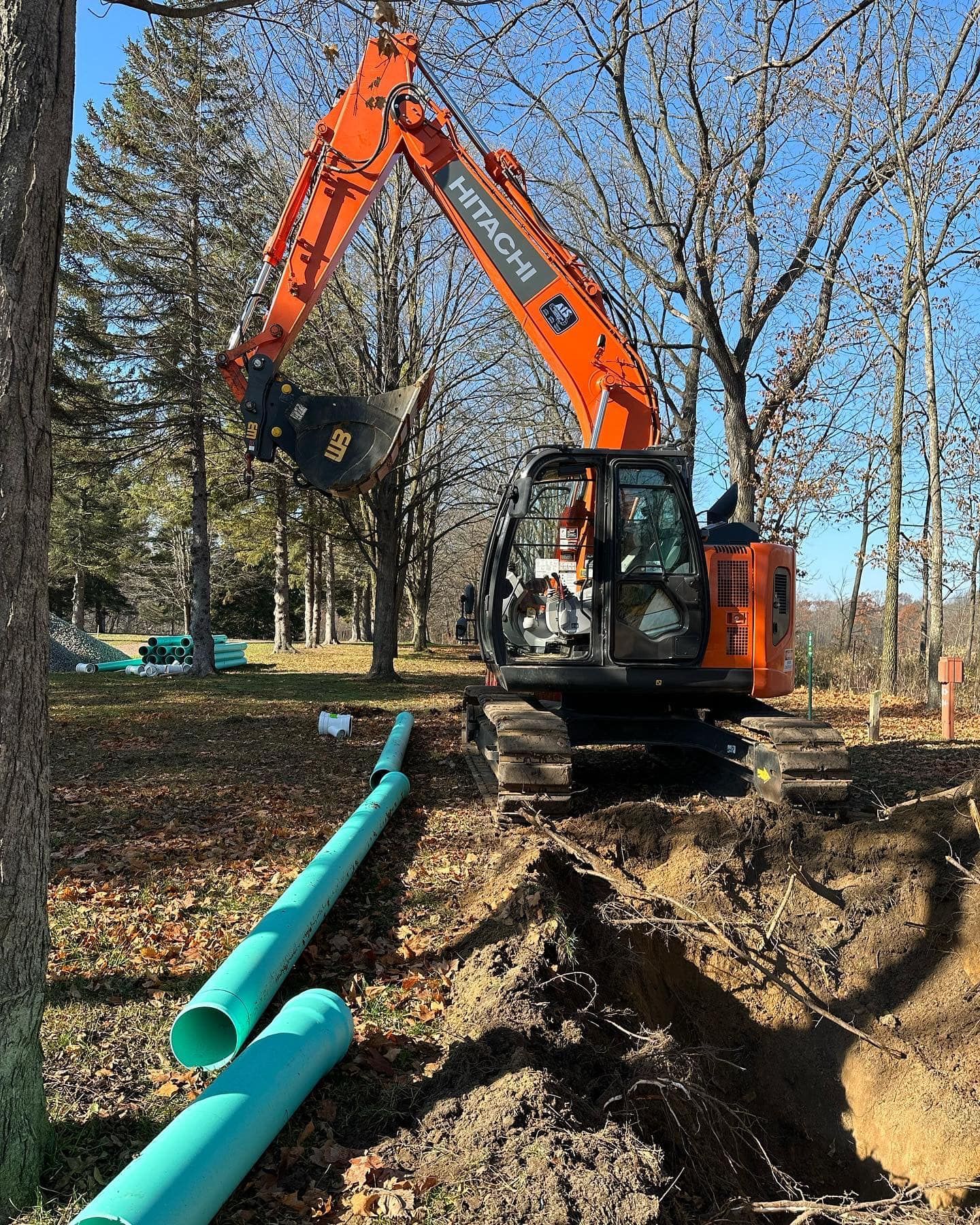 Orange Hitachi excavator digging a trench, with blue pipes nearby, in a wooded area.