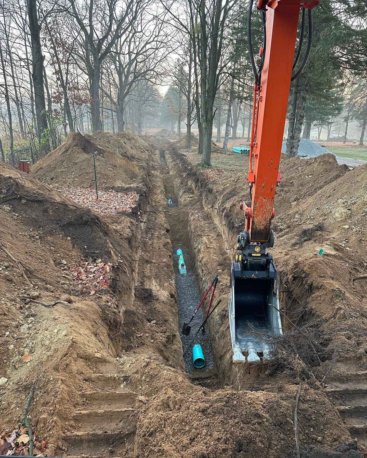An orange excavator digging a trench, with buried blue pipes in it, set in a wooded area.