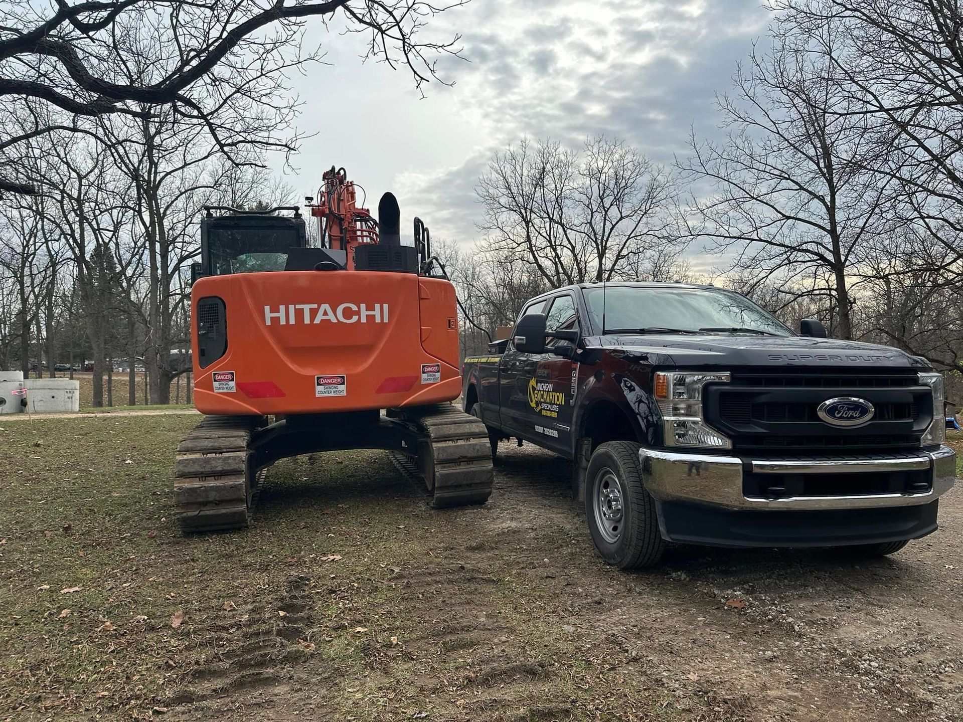 Orange Hitachi excavator next to a black Ford pickup truck in a grassy area with trees.