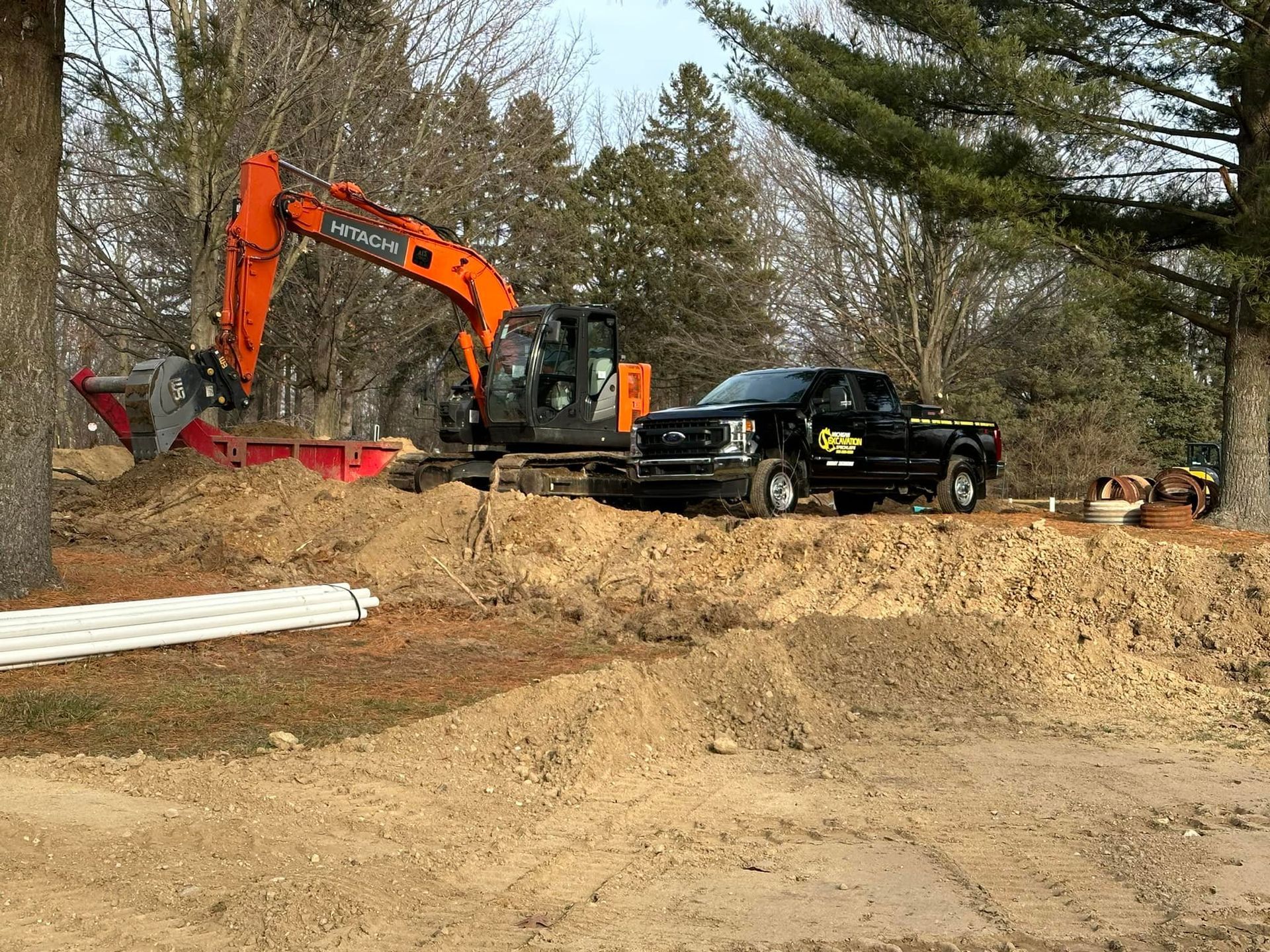 An excavator and black pickup truck on a mound of dirt. PVC pipes in the foreground.