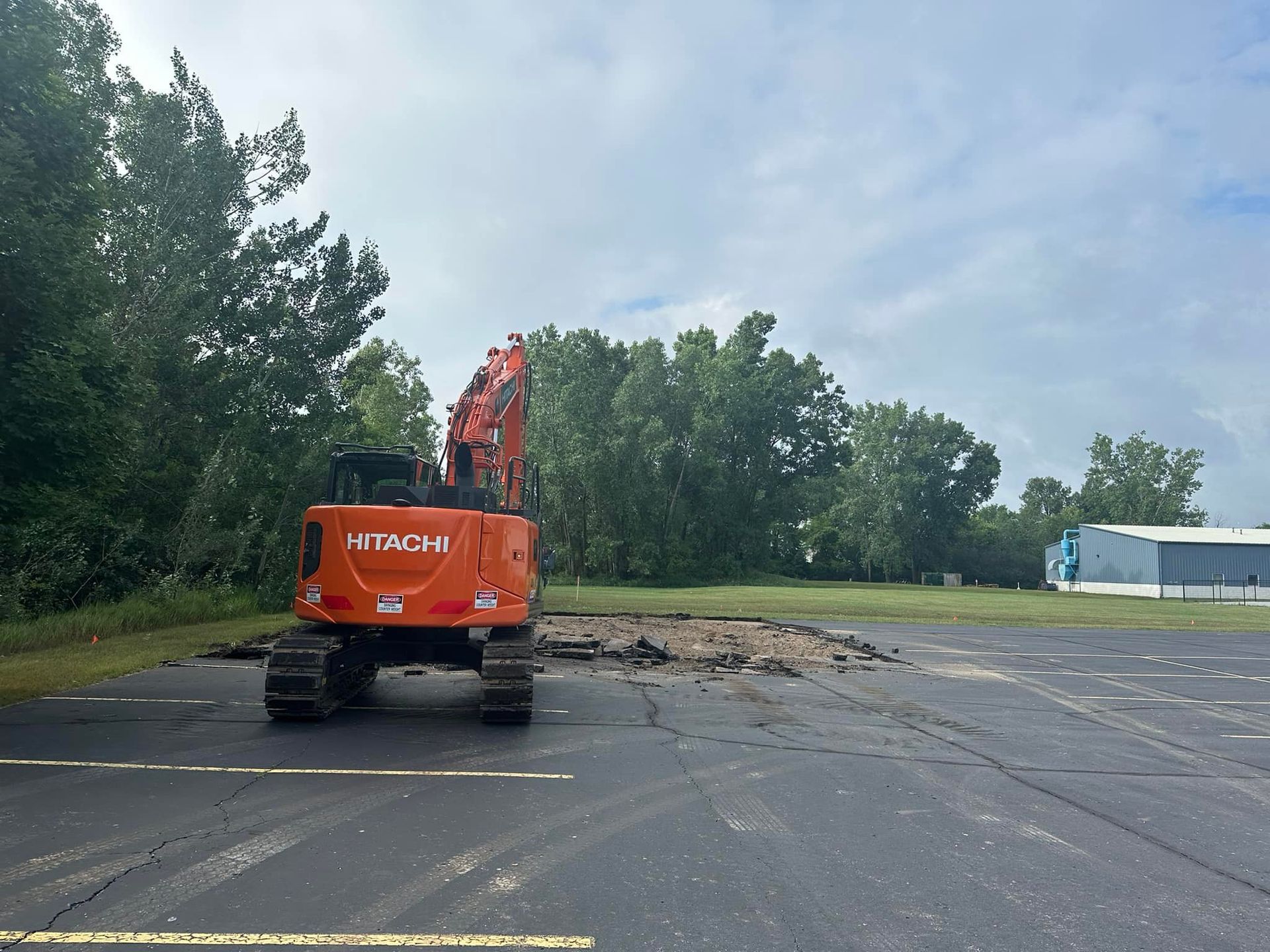 Orange excavator removing asphalt from a paved area with trees and a cloudy sky in the background.