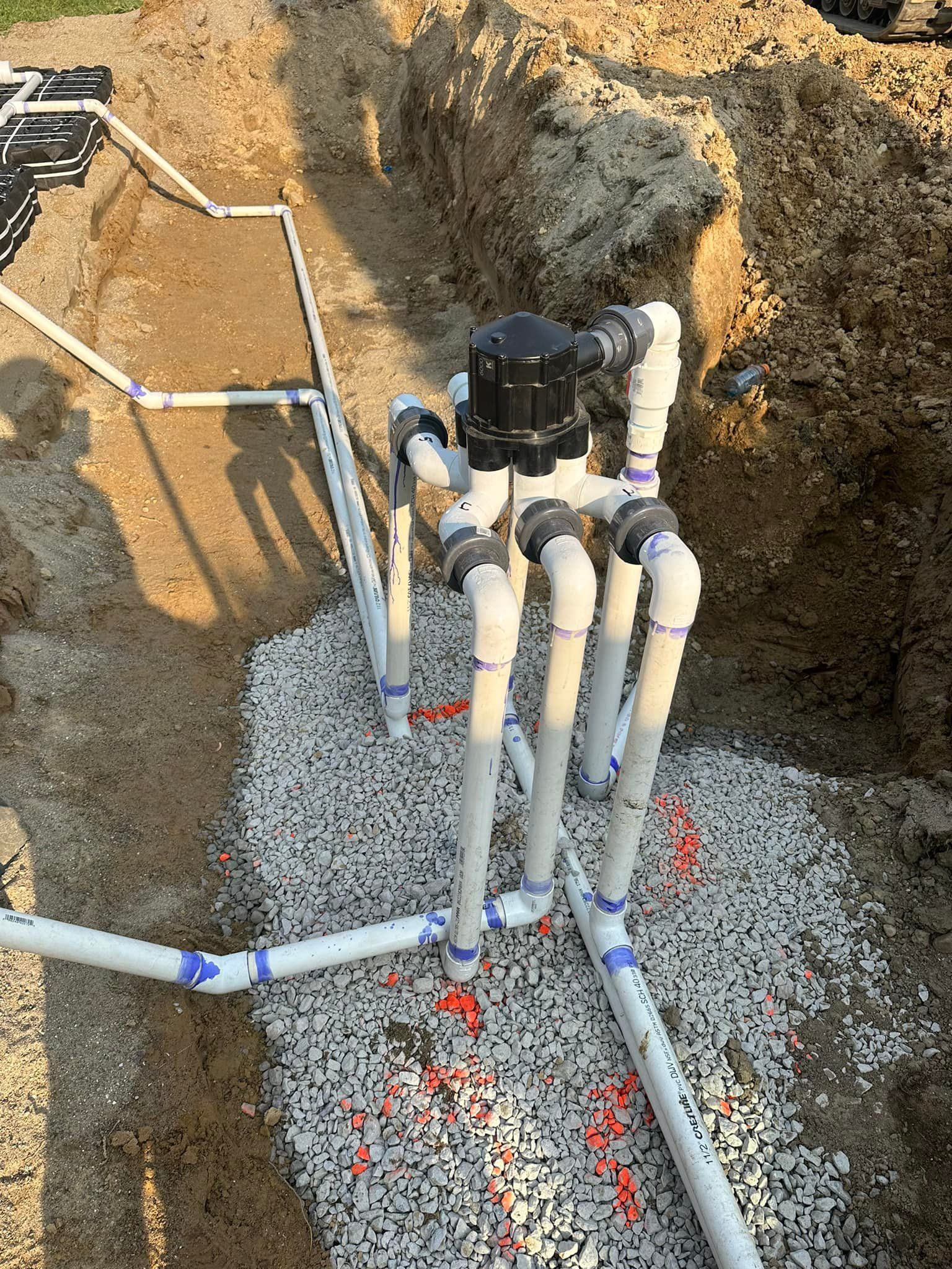 Pipes and a black valve system set in gravel within an excavated ditch. White PVC pipes and orange sprinkler heads.