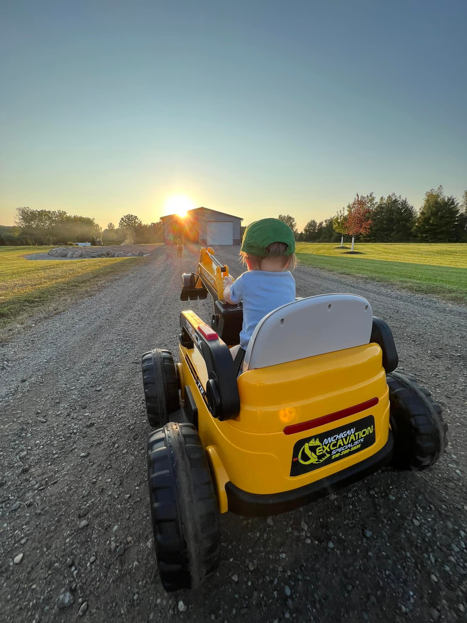 Child in a yellow toy digger driving on a gravel road toward the sunrise.