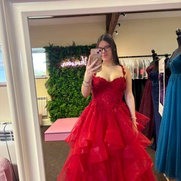 Woman in red formal dress taking a mirror selfie in a dress shop.