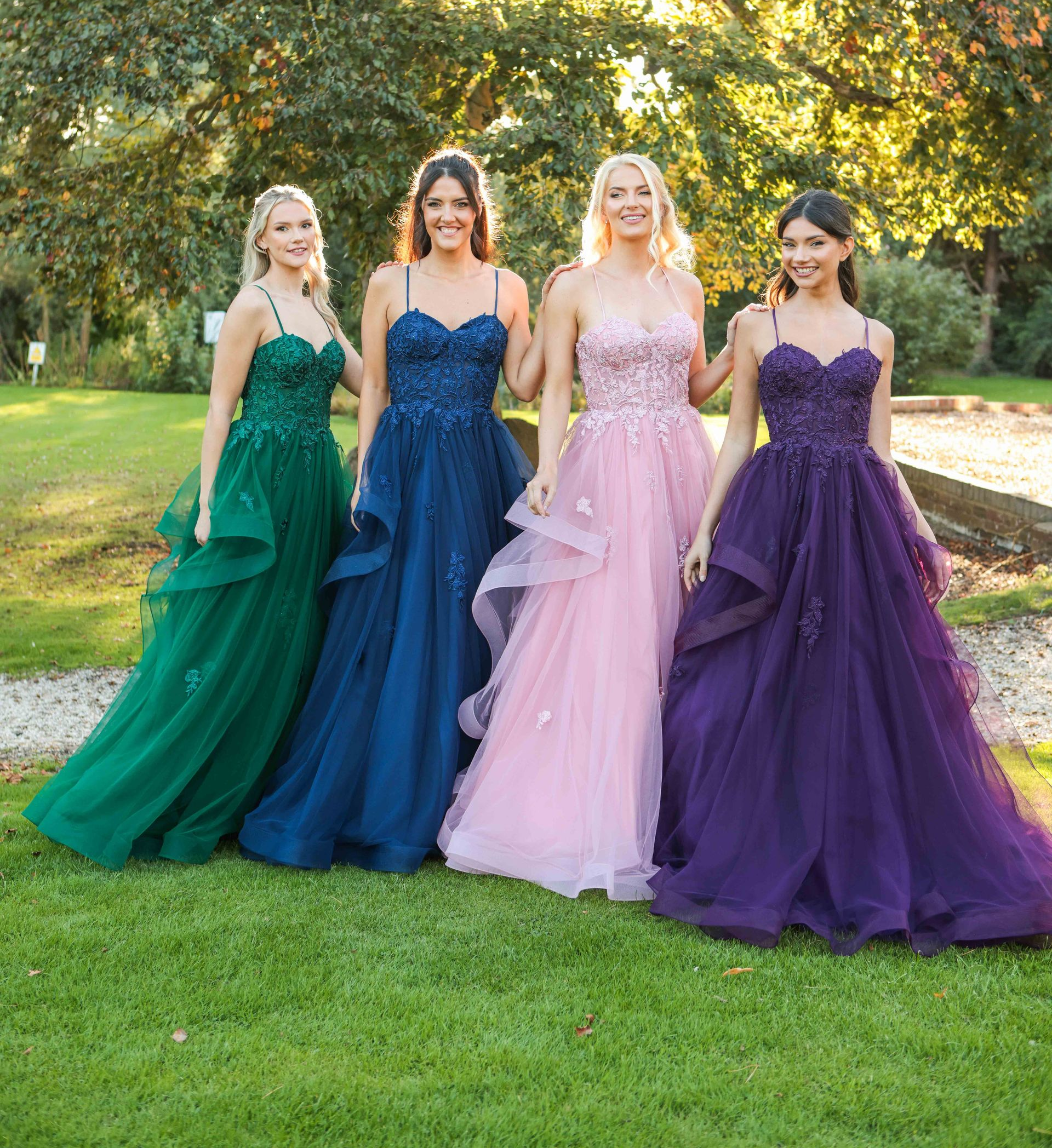 Four women in formal gowns posing outdoors: green, blue, pink, and purple dresses.