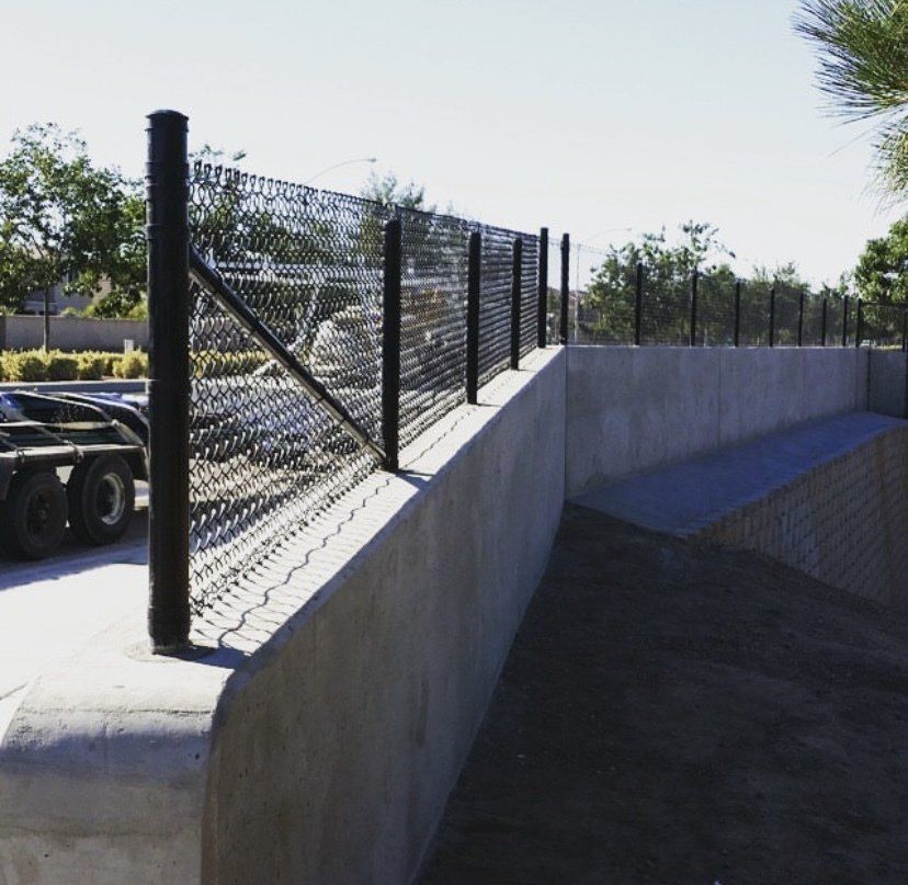 Chain-link fence atop a concrete wall, black posts, sunny day, trees in the background.