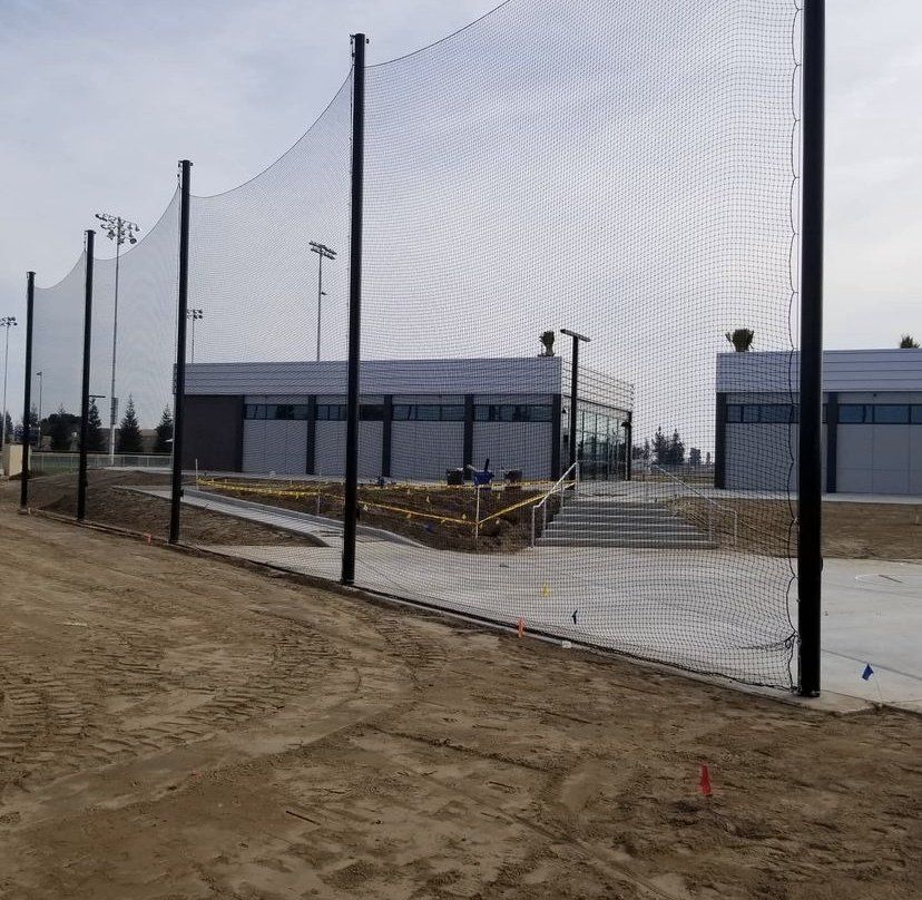 Netted structure and buildings on a dirt lot. Black poles support the netting. Gray buildings are in the background.