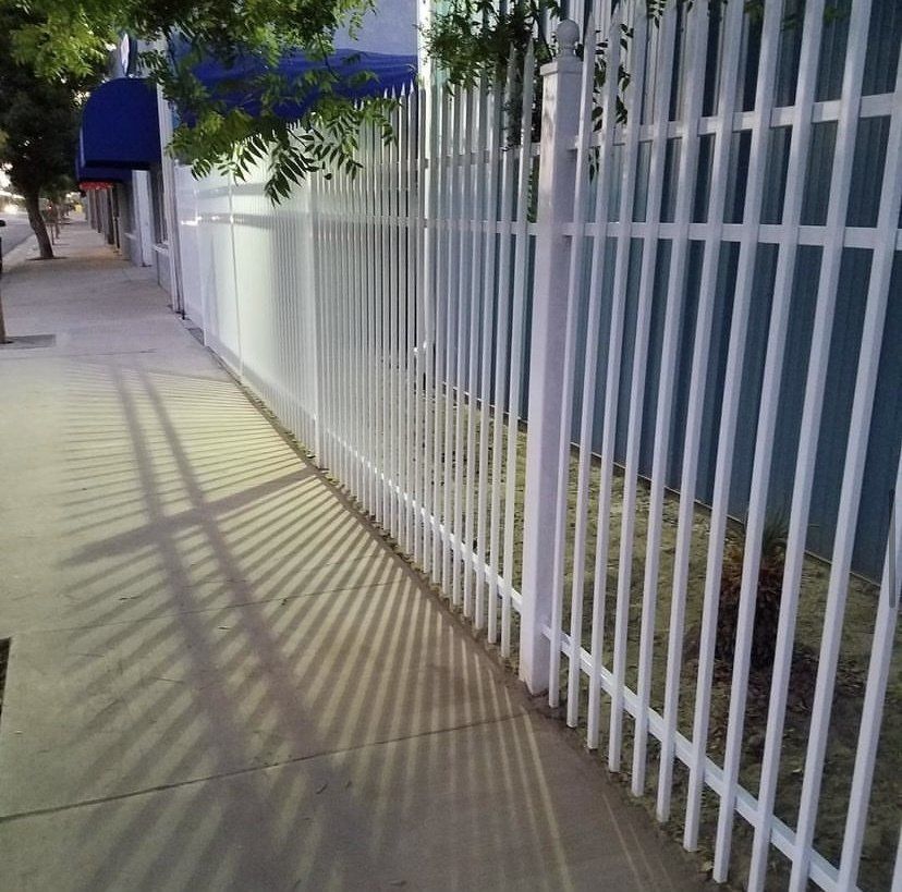 White metal fence casting long shadows on a sidewalk along a building.