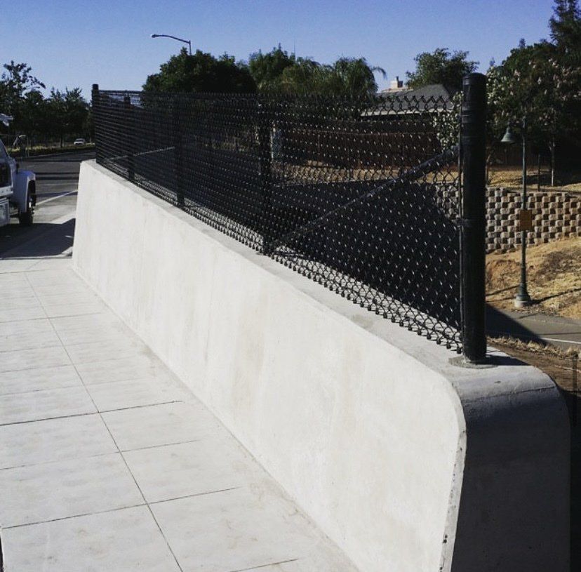 Black chain-link fence atop a concrete retaining wall on a sidewalk.