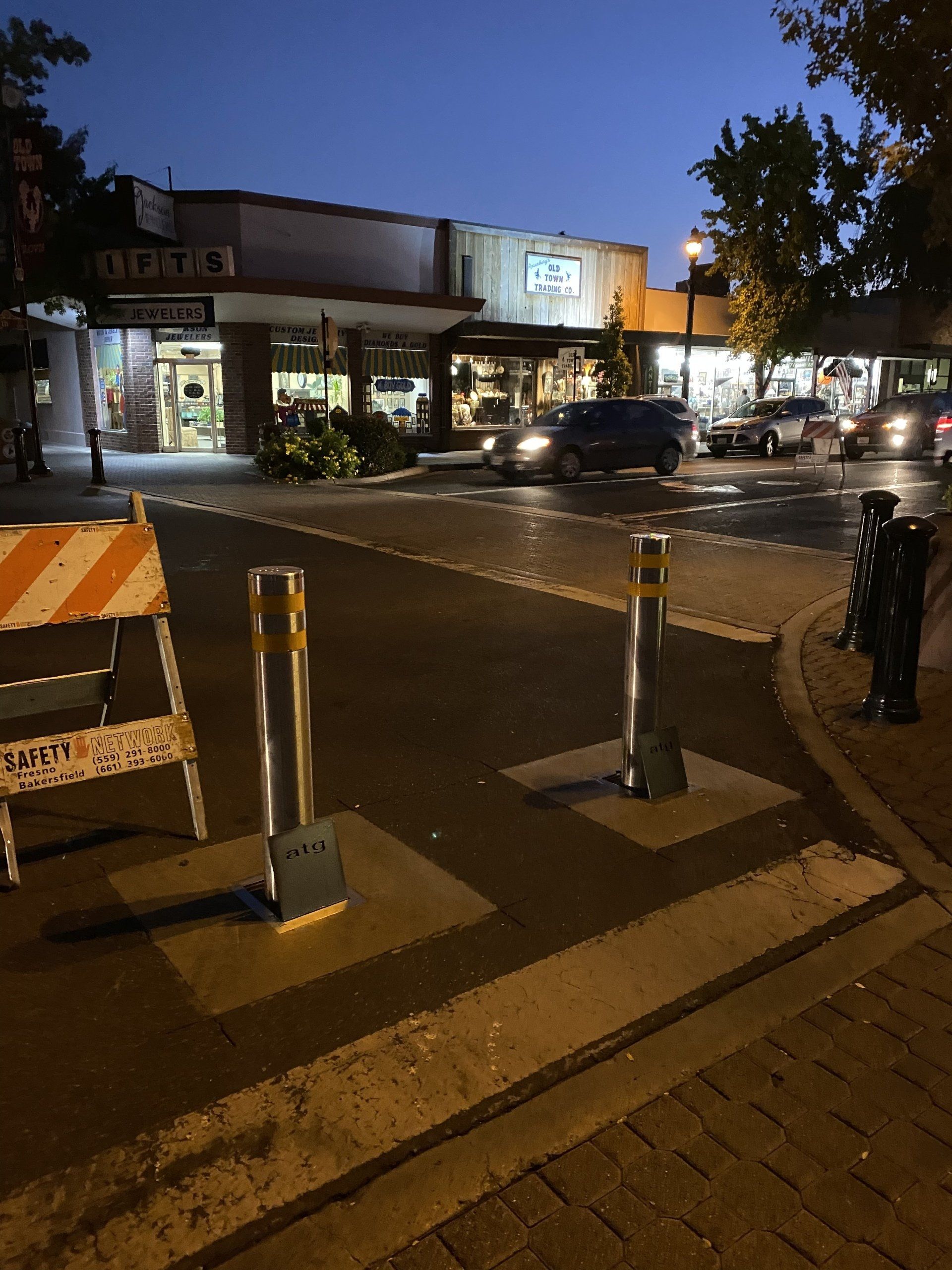 Street view at dusk with parked cars, businesses, and metal bollards.
