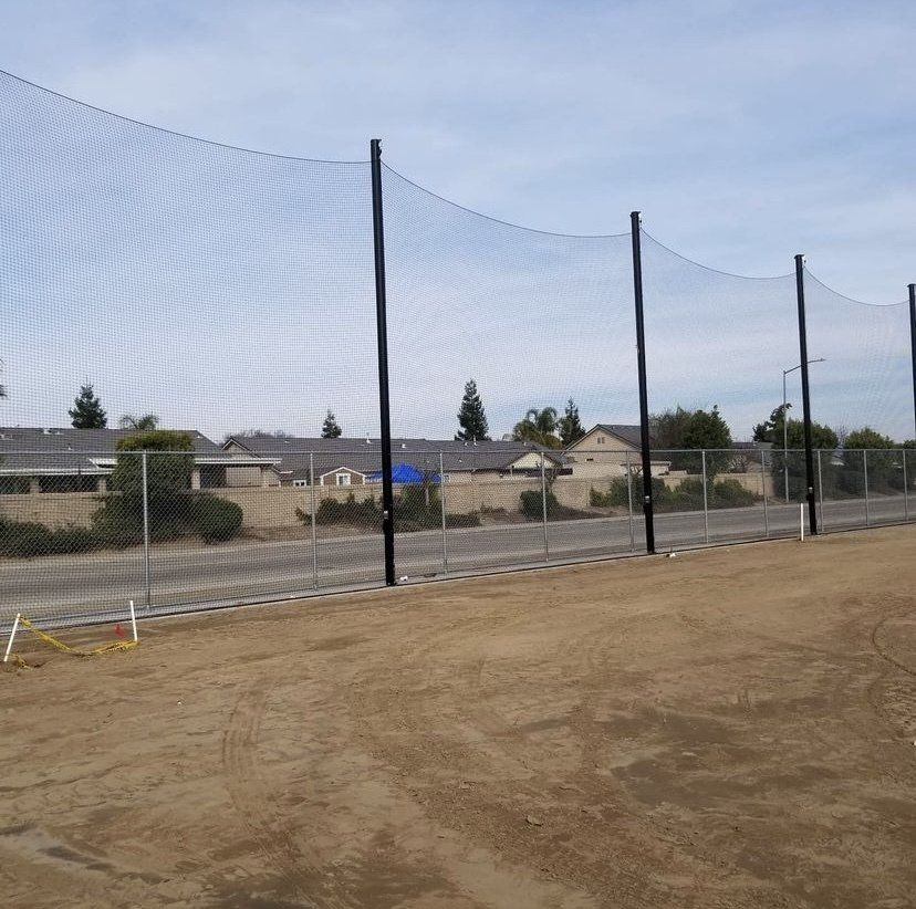 Large black netting on tall posts, near a chain-link fence, protects a dirt field. Homes are in the background.