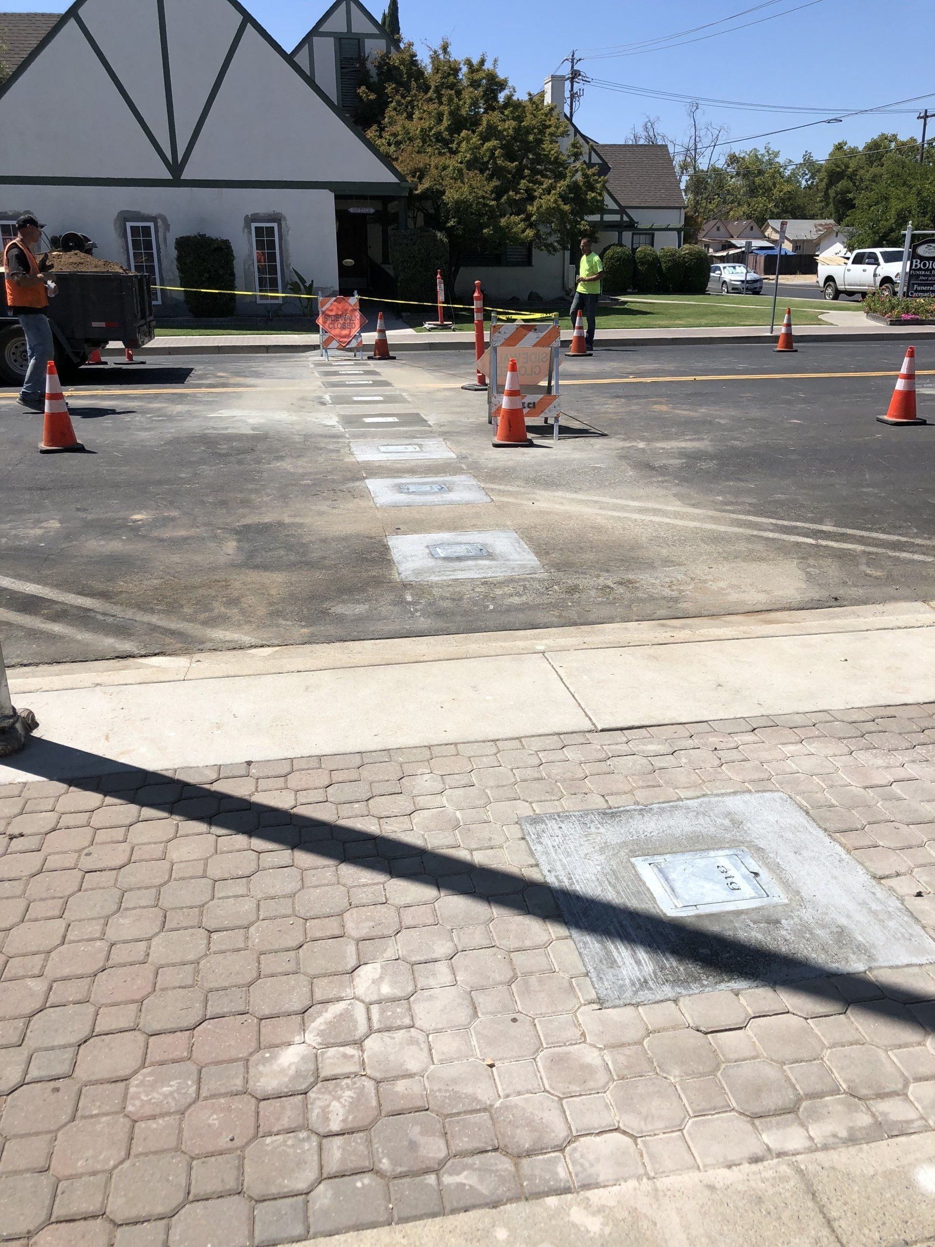 Road work in progress; open utility access covers in asphalt. Orange cones, workers visible, sunny outdoor setting.