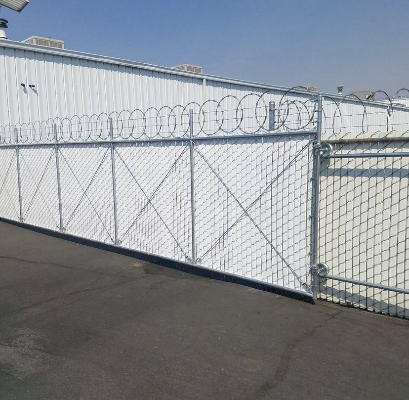 Chain-link fence topped with razor wire. Gray metal, white building in background, asphalt ground, sunny day.