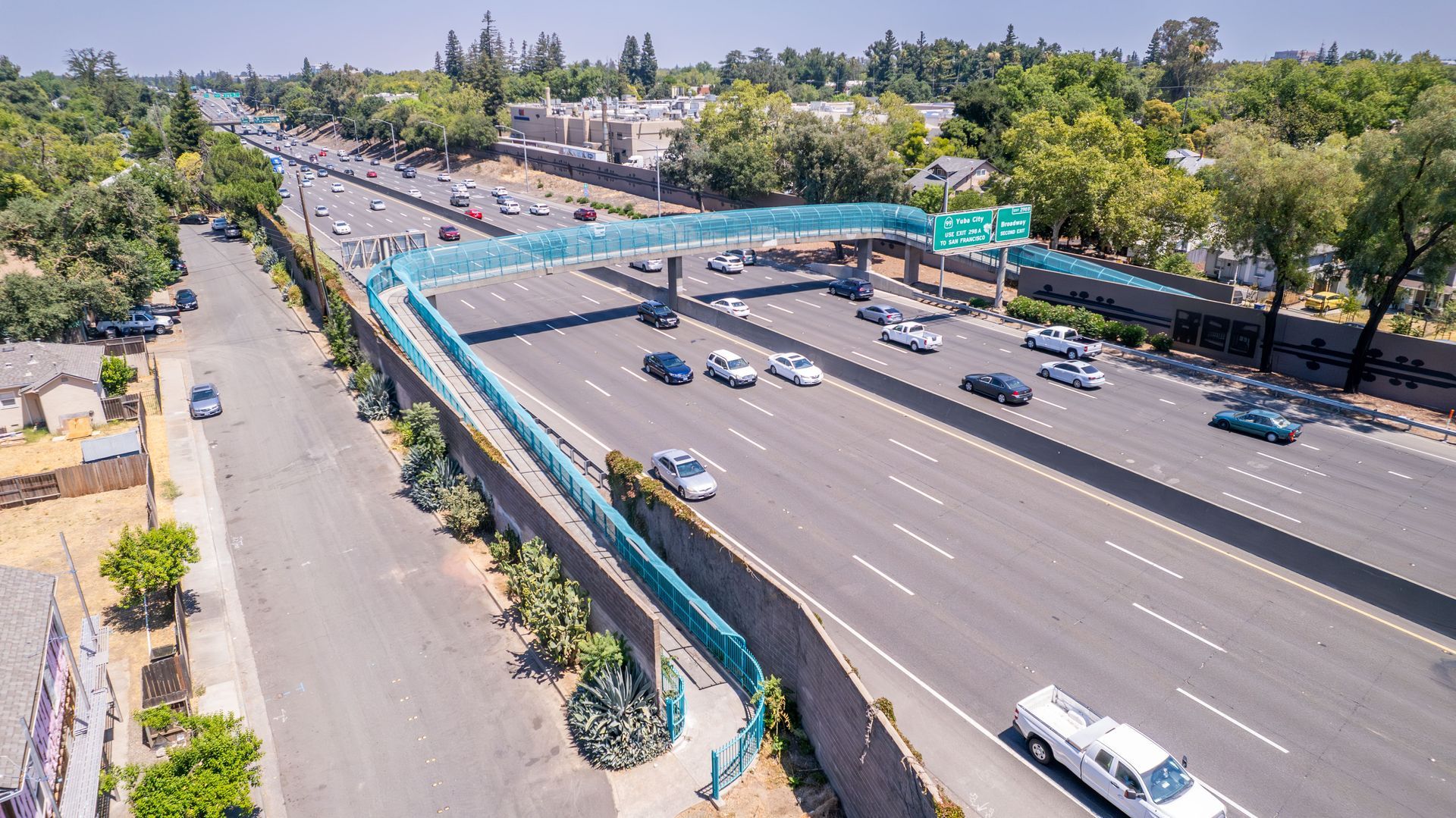 Aerial view of highway with pedestrian bridge. Cars travel on the highway. Bridge is blue, with green signage.