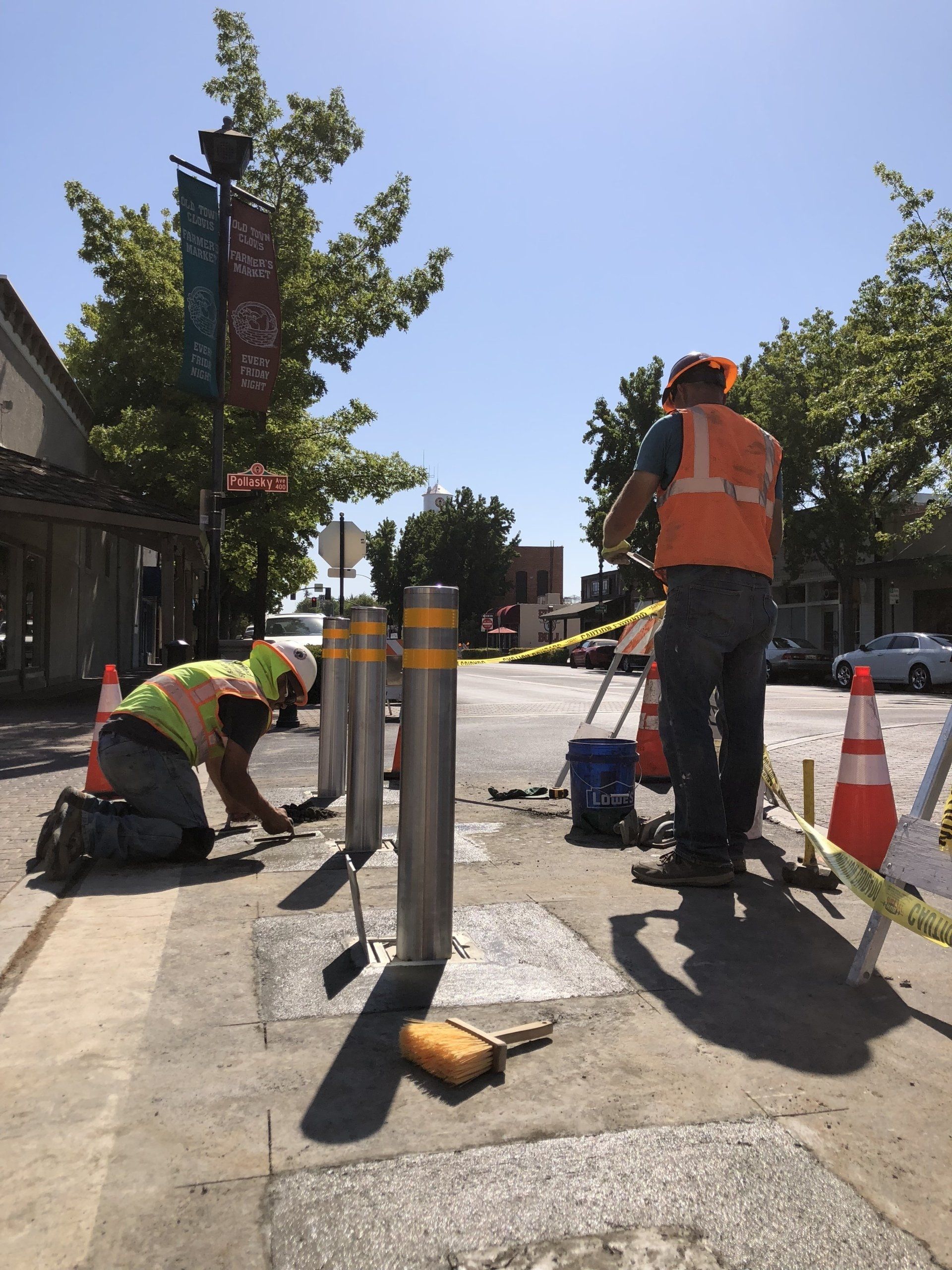 Construction workers install bollards on a sidewalk under a sunny sky.