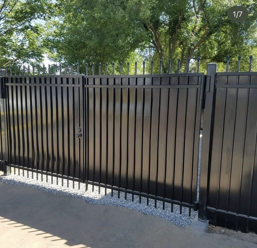 Black metal gate with vertical bars and a lock, set against a backdrop of trees and gravel.