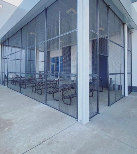 Outdoor seating area enclosed by a metal mesh fence. Tables and benches visible.