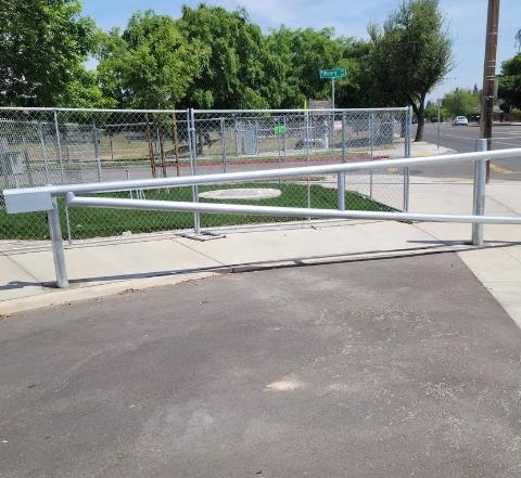 A metal ramp, gray fence, and green grass are in front of a street with trees under a blue sky.