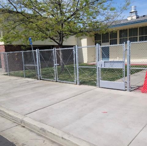 Chain link fence with gates in front of a building on a sidewalk.