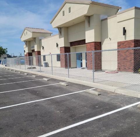 Exterior view of a beige building with a chain-link fence surrounding a parking lot. Blue sky overhead.