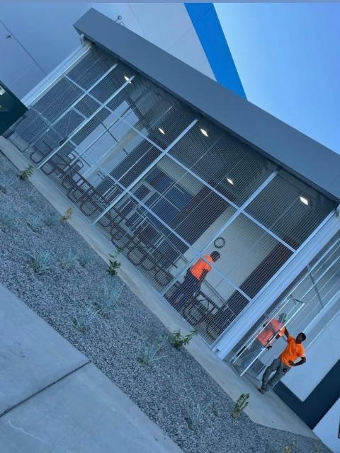 Two workers in orange vests near a building with glass windows and a bicycle rack. Outdoors.