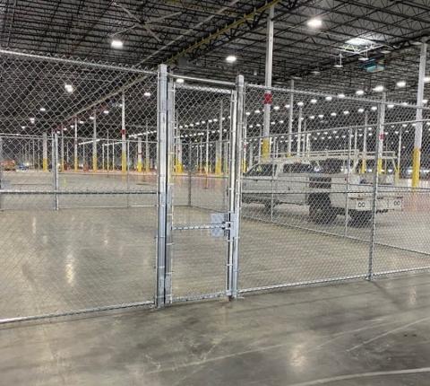 Chain-link fence and gate inside a large warehouse, possibly for secure storage. A truck is visible in the background.