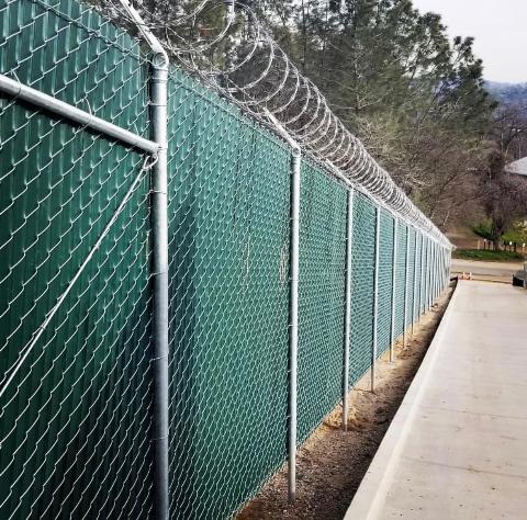 Green chain-link fence with green privacy slats and razor wire along the top, outdoor setting.