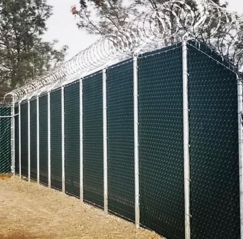 Dark green chain-link fence with privacy slats and razor wire topper, set in a dirt lot.