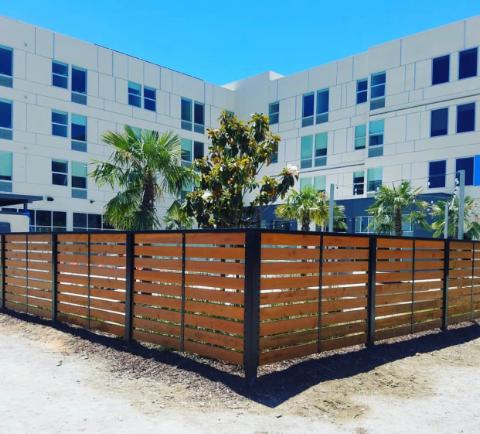 Wooden slat fence in front of a multi-story building with palm trees and a blue sky.