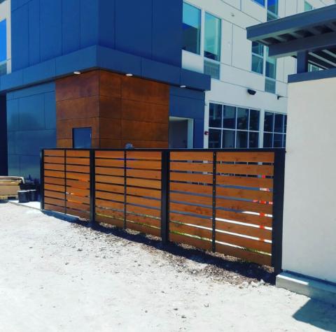 Wooden slat fence with black metal frame in front of a modern building.