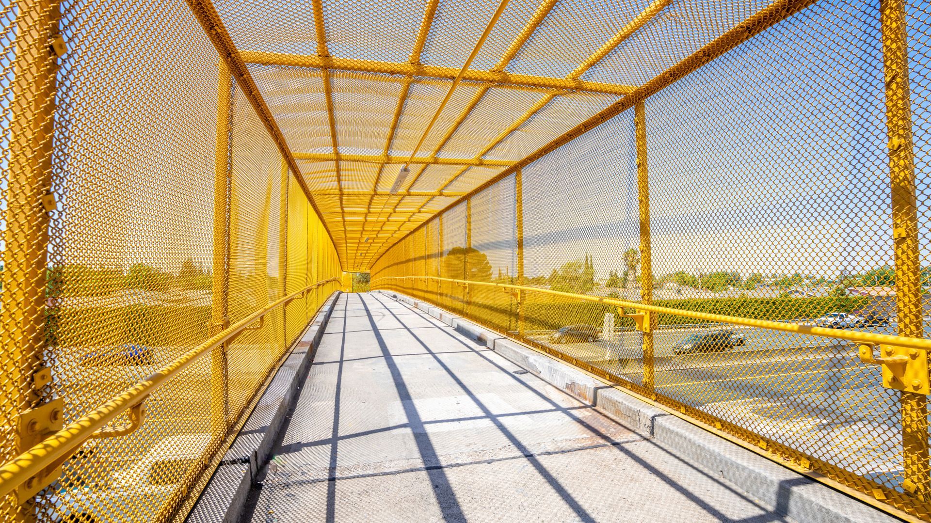 Yellow chain-link pedestrian bridge, concrete path, enclosed with railings, casting shadows in the bright sunlight.