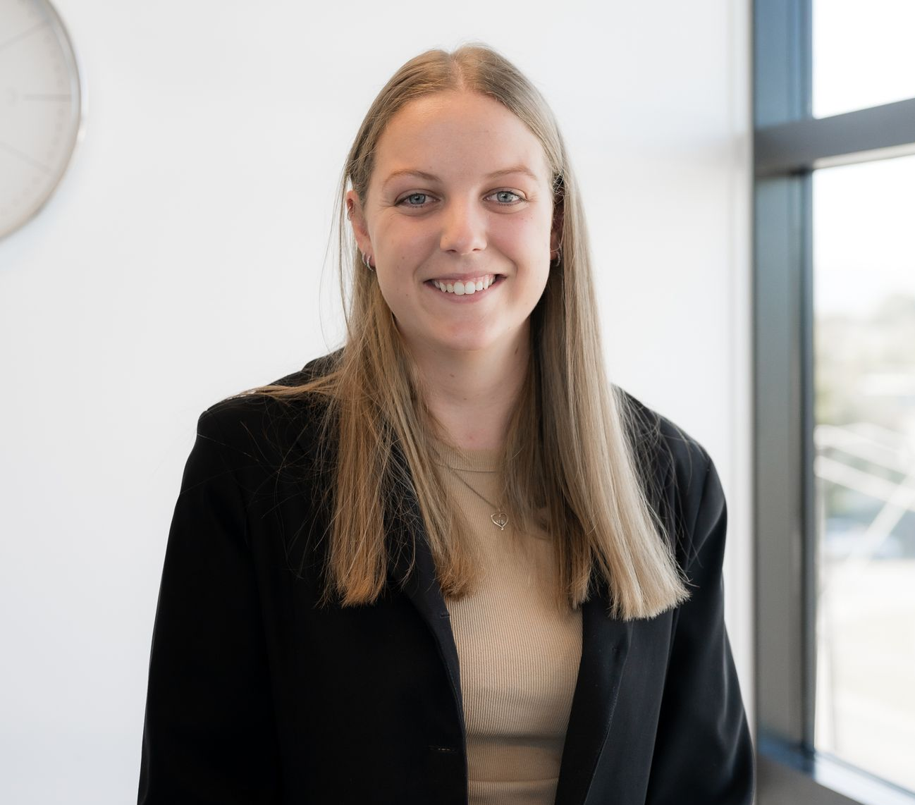 A smiling person with long light brown hair wears a black blazer over a tan ribbed top in a bright, modern office.