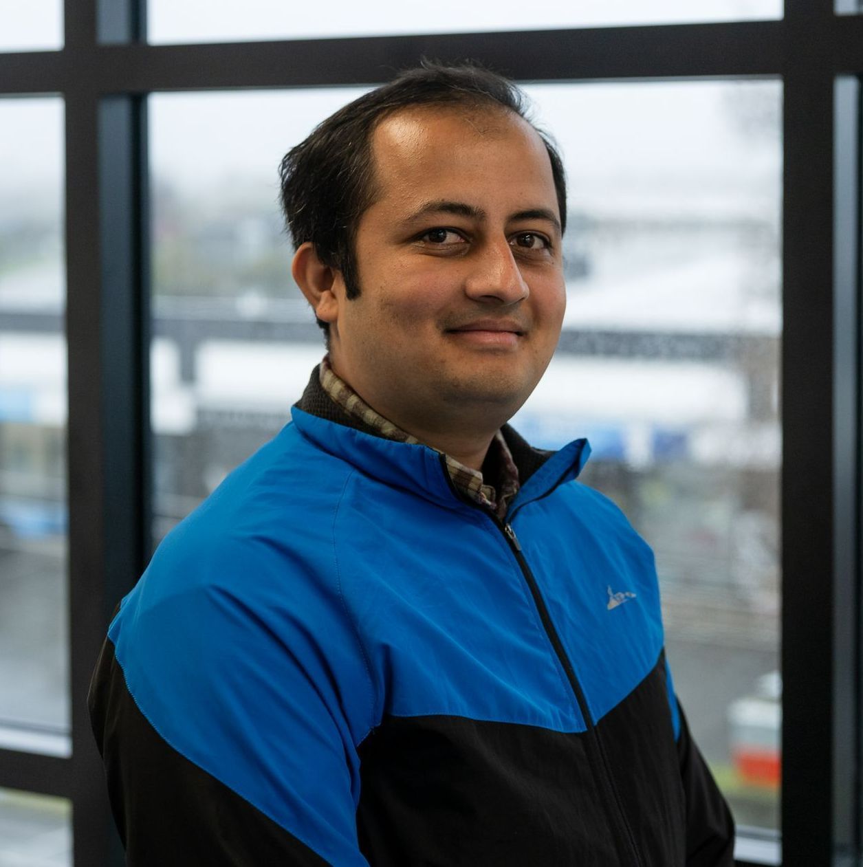 Prabin Kattel in a blue and black jacket is standing in front of a window .