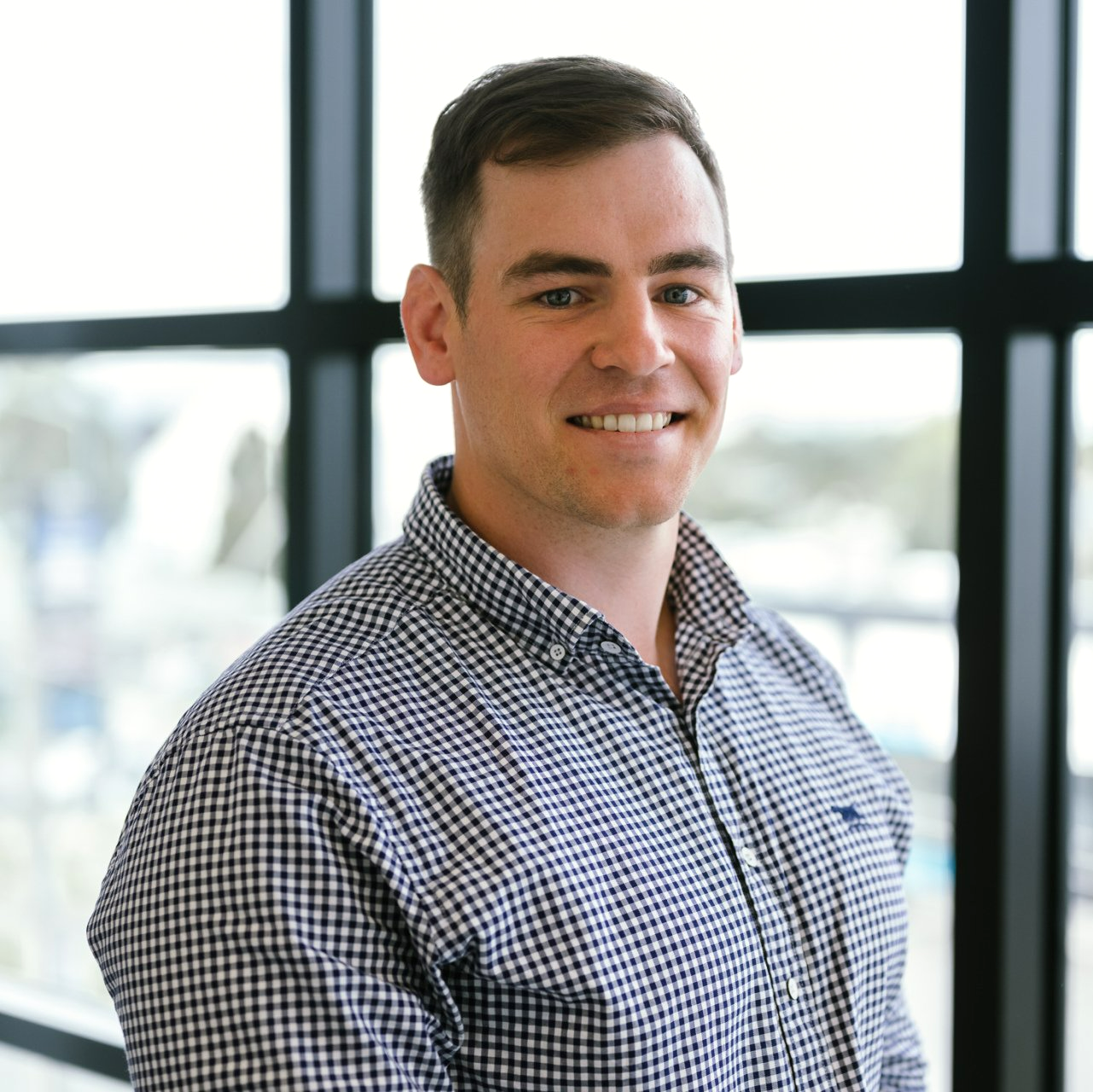 Sam Foster in a plaid shirt is smiling in front of a window .