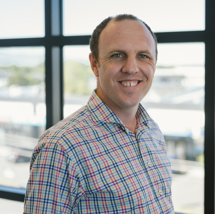 A man in a plaid shirt is smiling in front of a window.