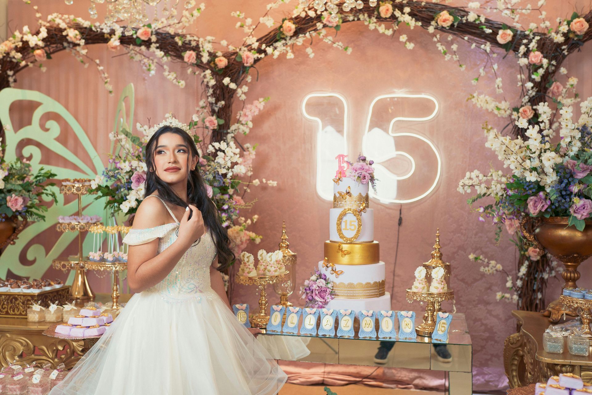 Girl in white dress posing beside a birthday cake and neon “15” at a pink floral celebration backdrop