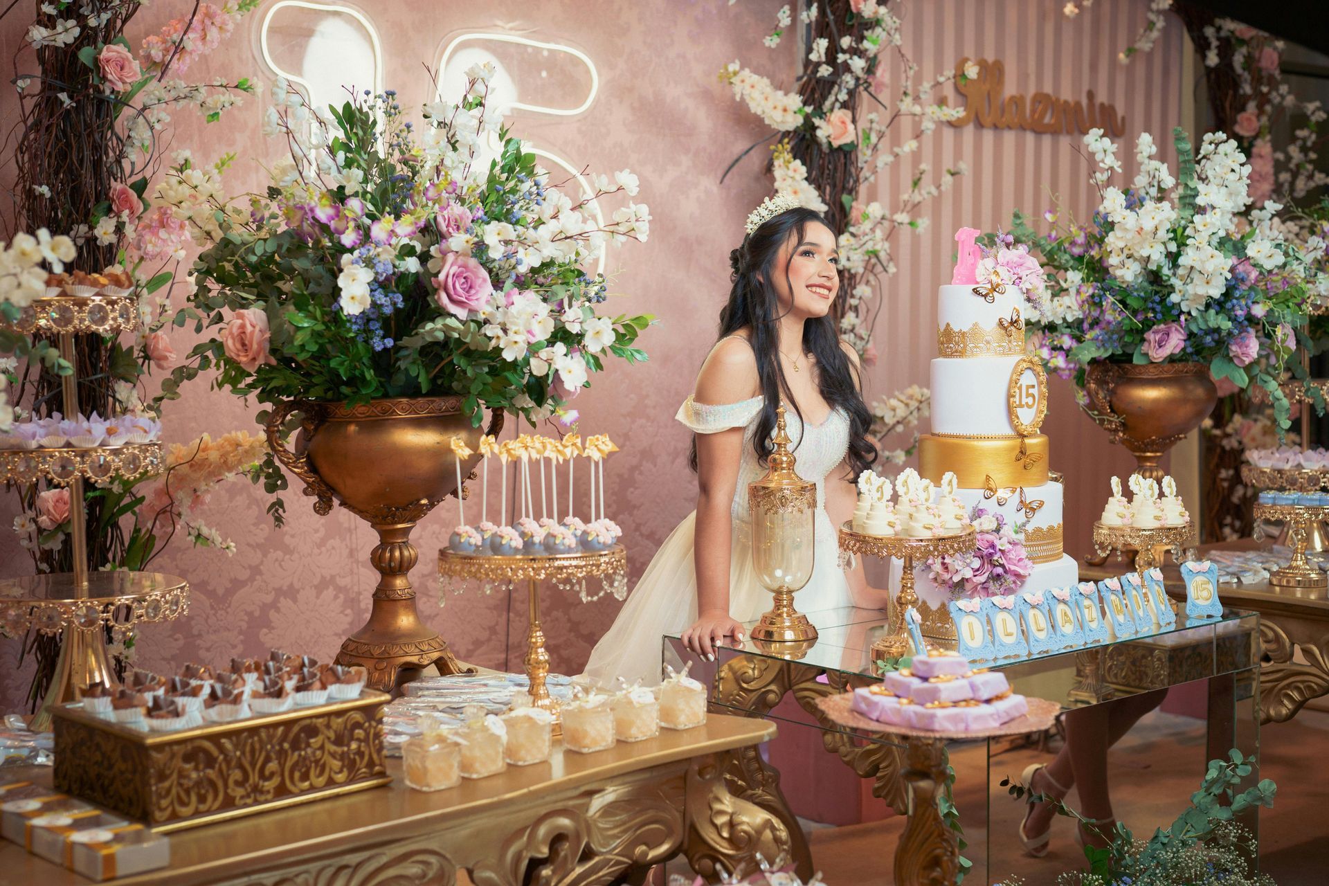 Elegant dessert table with pink flowers, gold decor, and a woman in a white dress at a lavish party backdrop