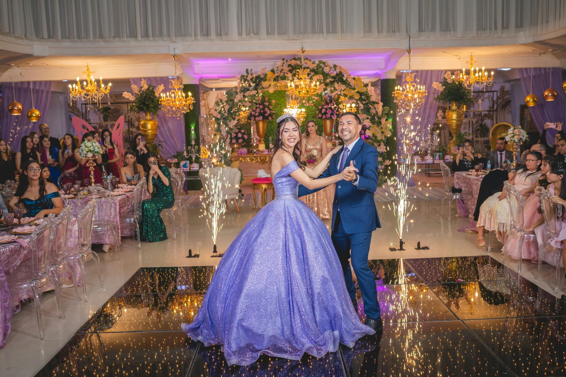 Couple dancing at a decorated ballroom celebration with purple lighting and guests seated nearby