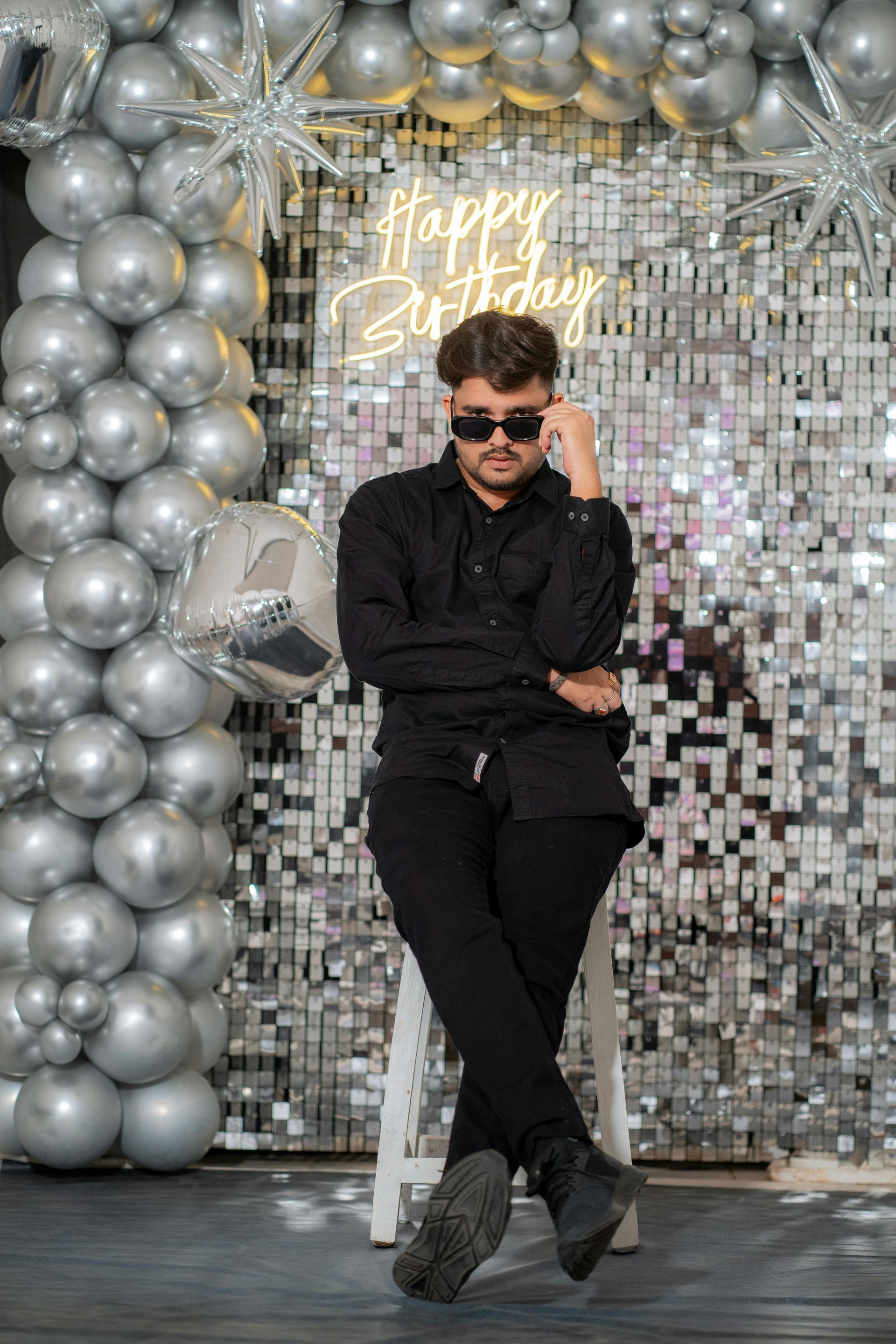 Person in black outfit standing before silver balloon-and-mirror birthday backdrop with “Happy Birthday” sign