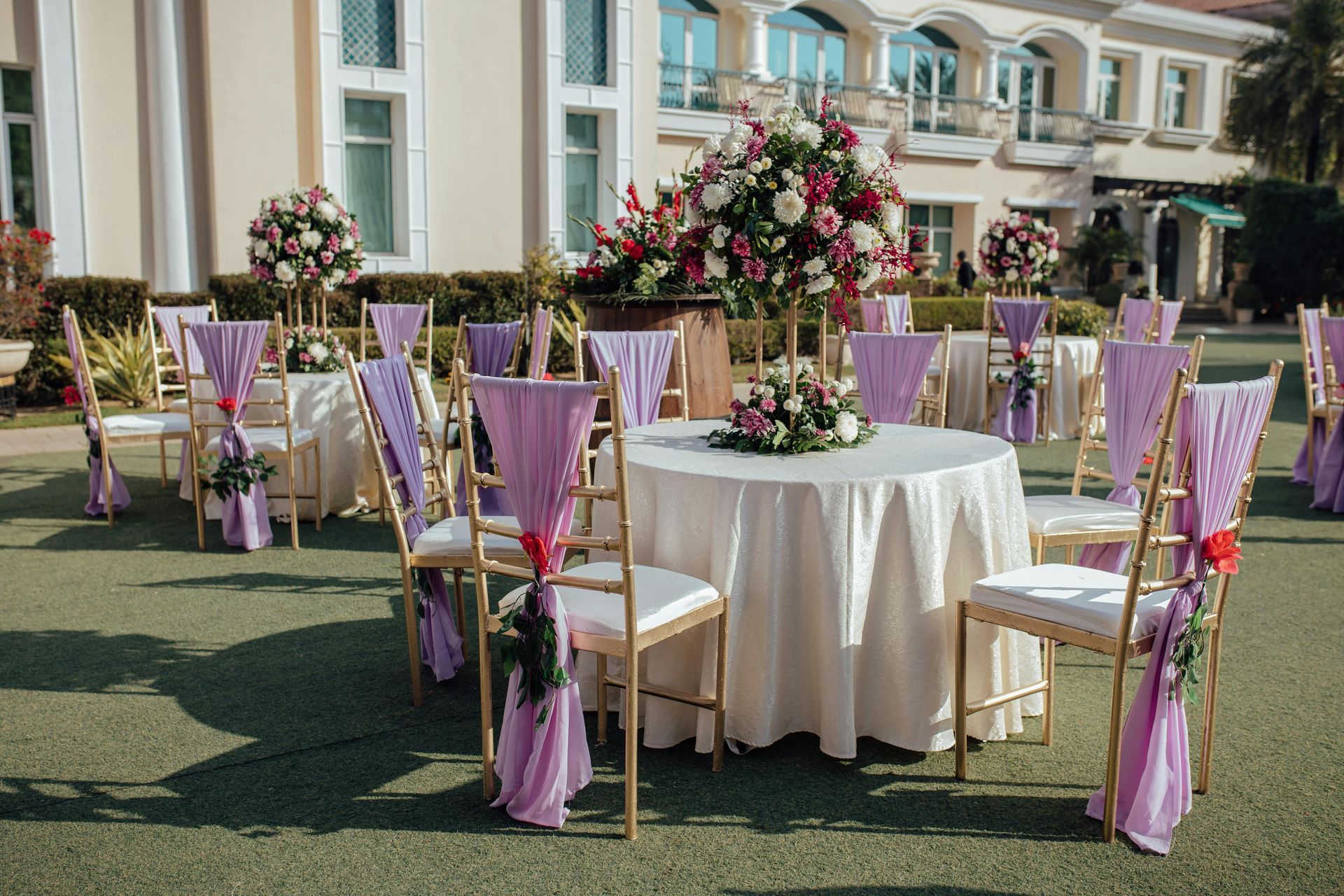 Decorated outdoor tables with floral centerpieces and lavender chairs on a lawn in front of a building