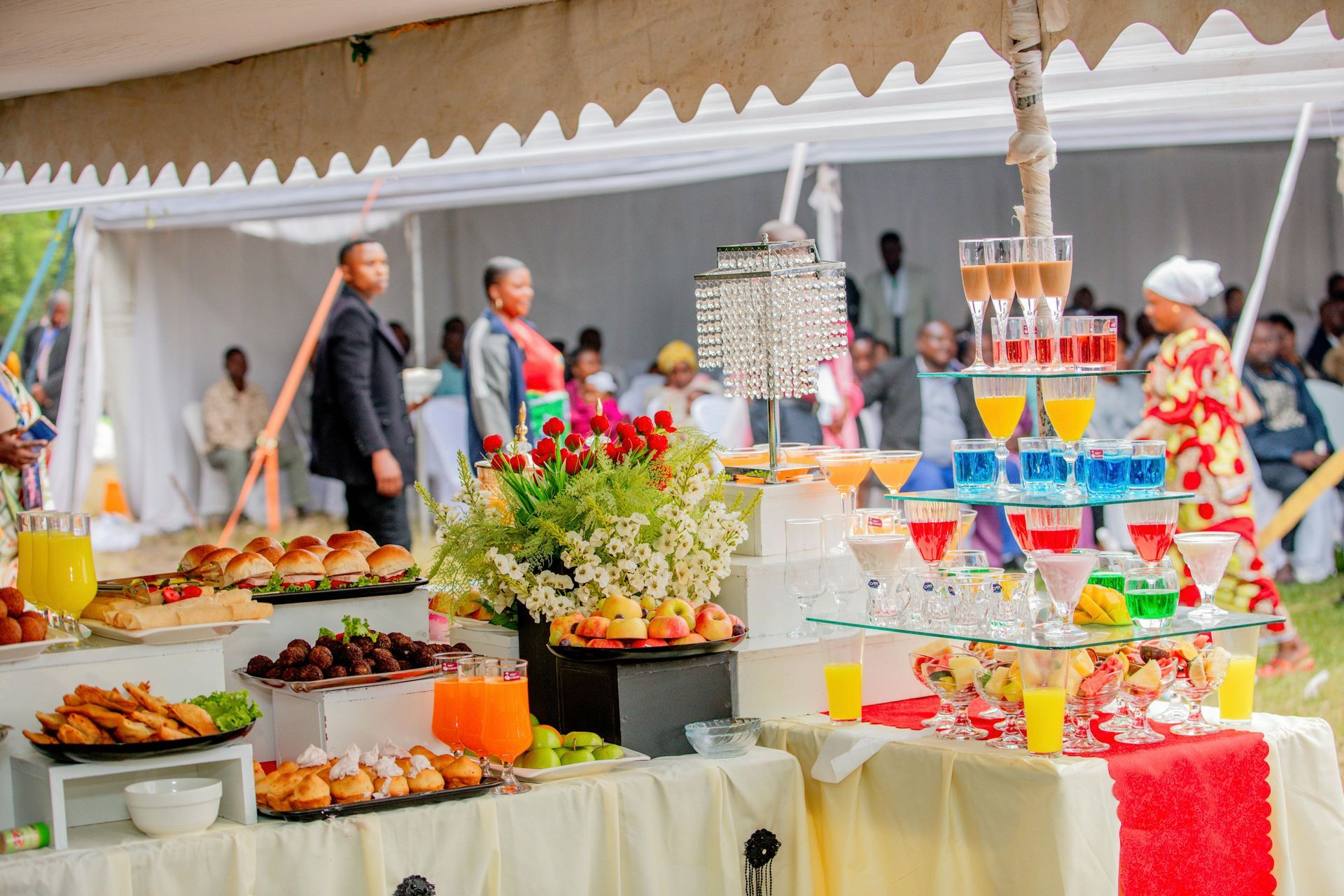 Outdoor buffet table with colorful desserts, drinks, and appetizers under a white tent