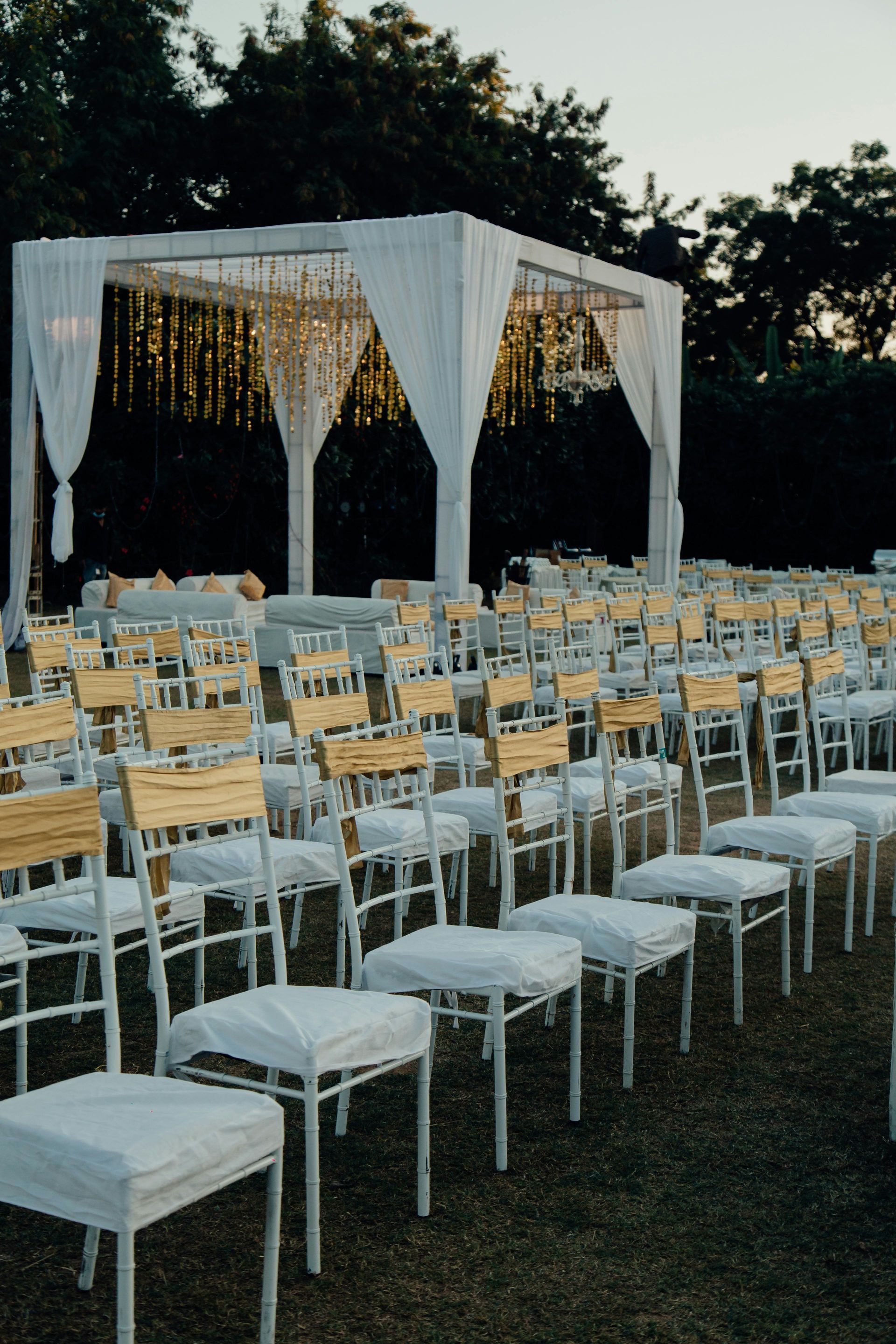 Outdoor wedding ceremony setup with rows of white chairs and a white draped canopy at dusk