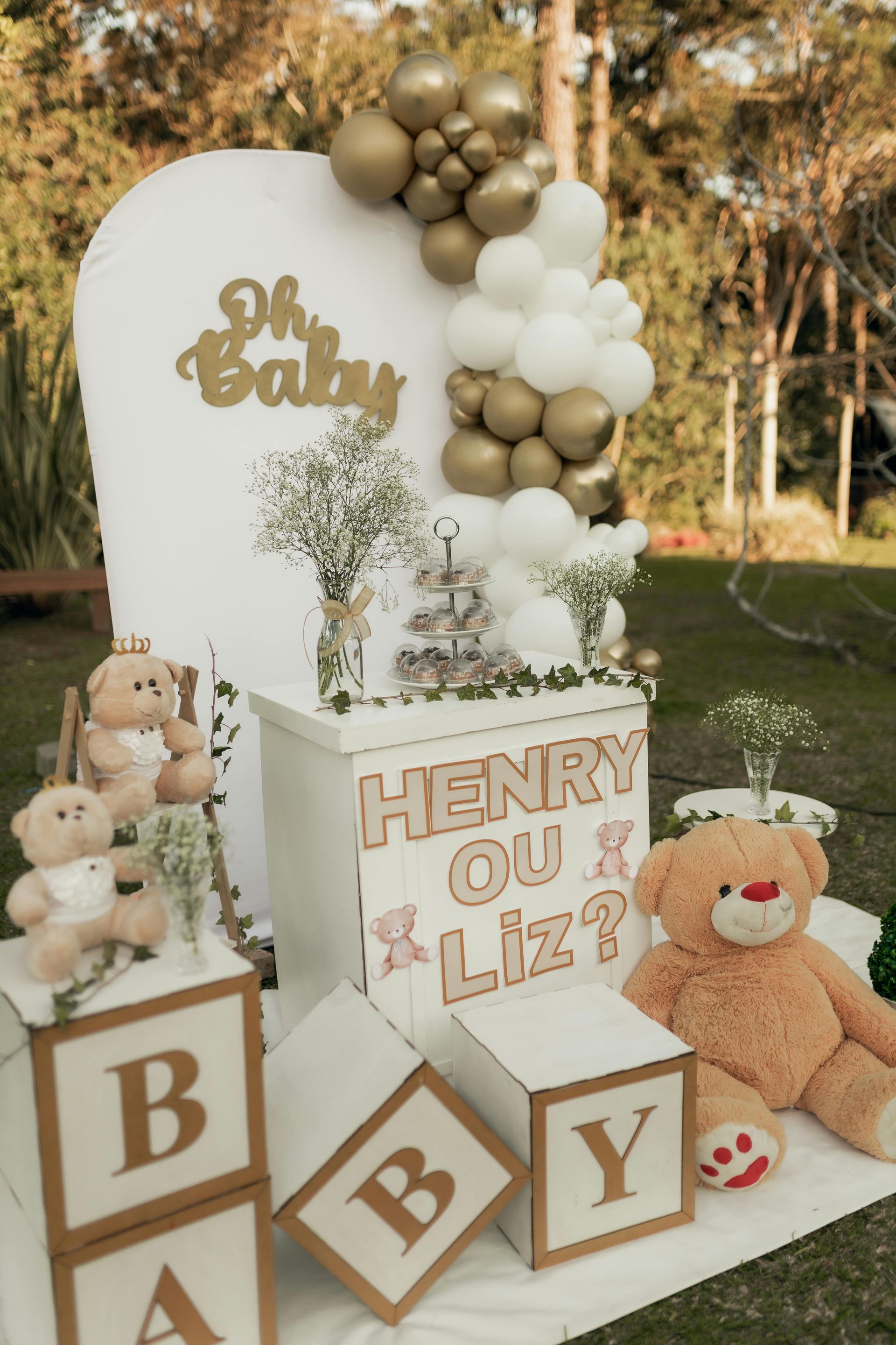 Baby shower display with white and gold balloons, teddy bears, and “HENRY OU LIZ?” blocks and banner