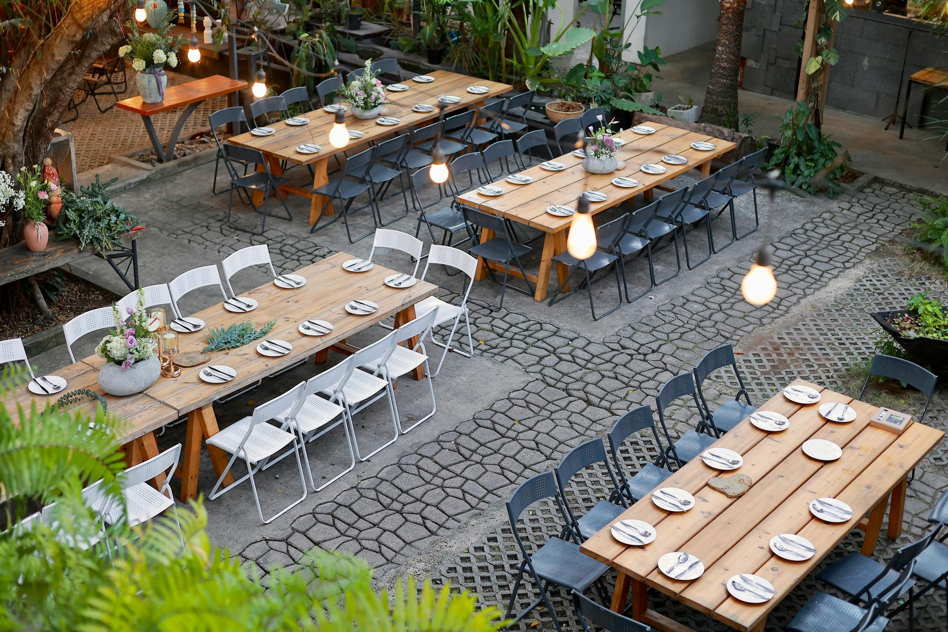 Outdoor patio with long wooden dining tables, white chairs, and hanging lights in a cobblestone courtyard