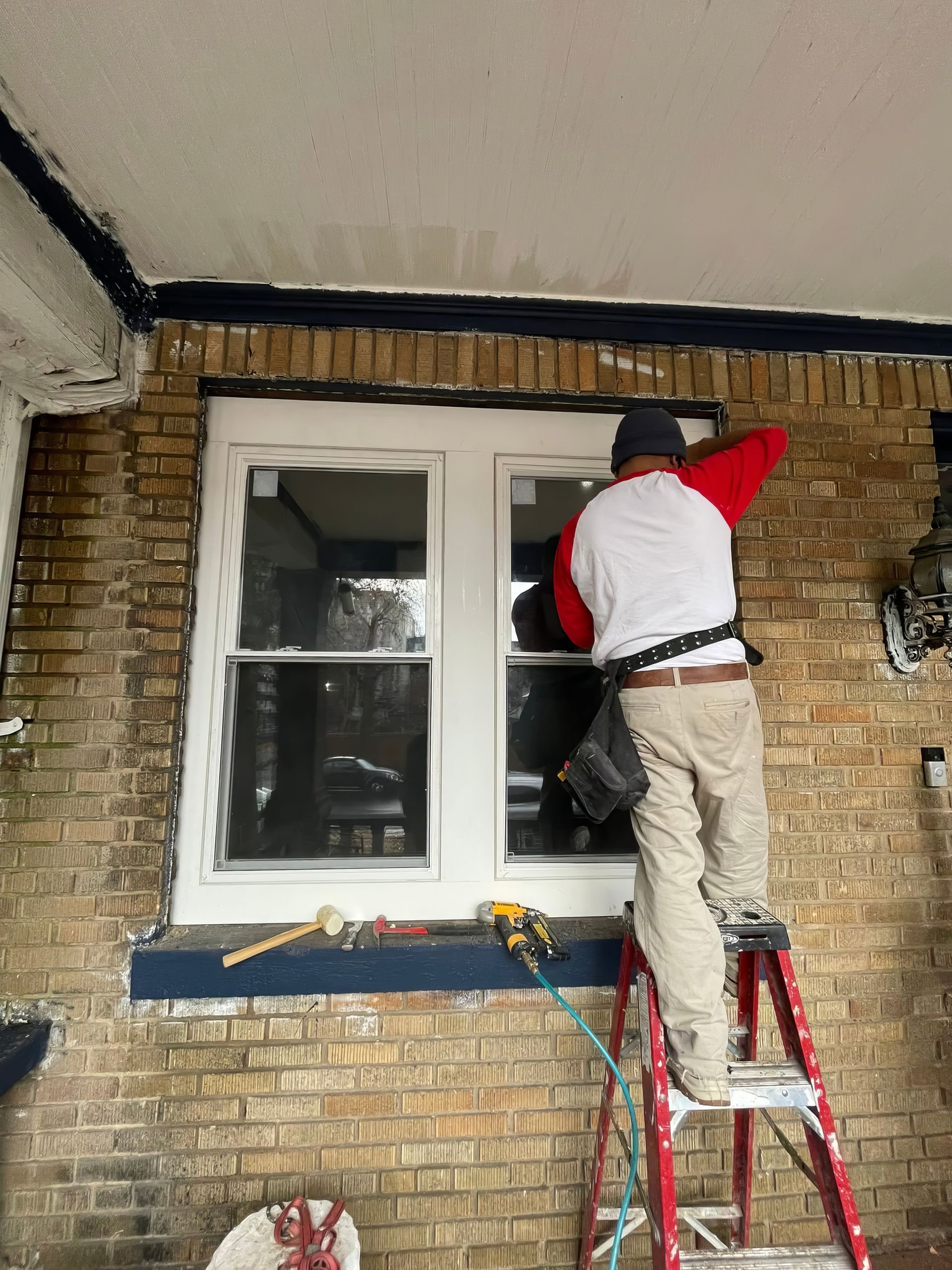 Worker on ladder painting the exterior wall above a white window on a brick house.