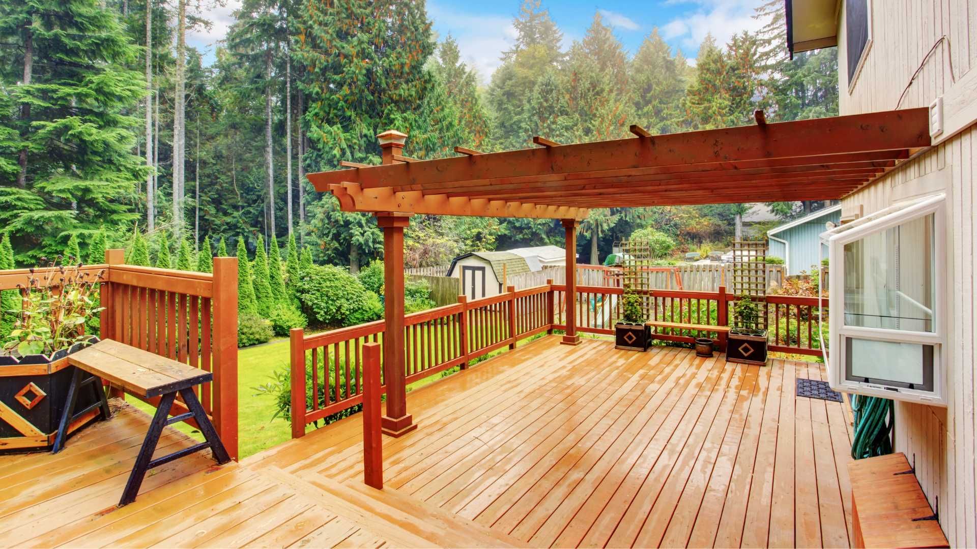 Wooden deck with pergola and railing overlooking a forest, with outdoor seating and a hot tub area