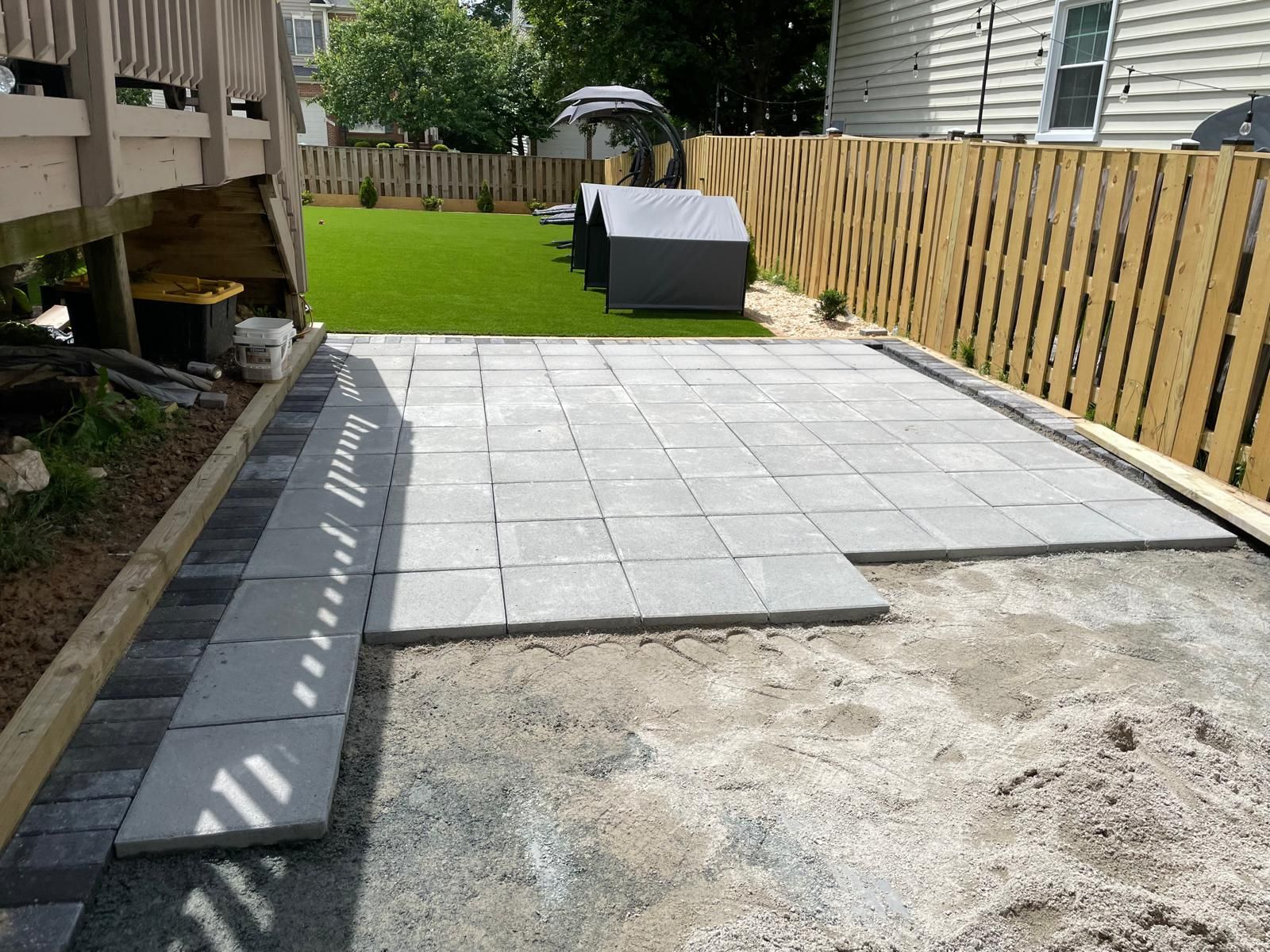Backyard patio with newly laid gray pavers beside a wooden fence and green lawn.