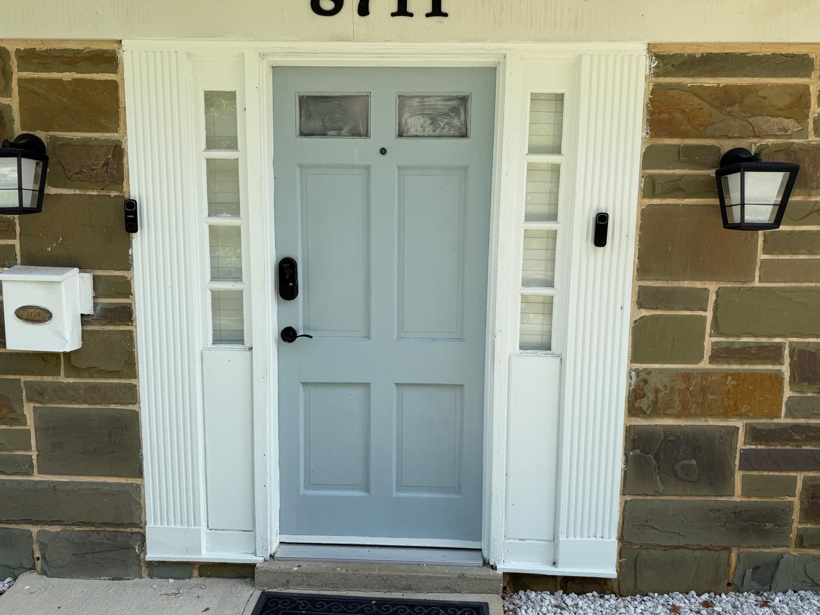 Front entrance with light gray door, stone exterior, black wall lanterns, and white trim