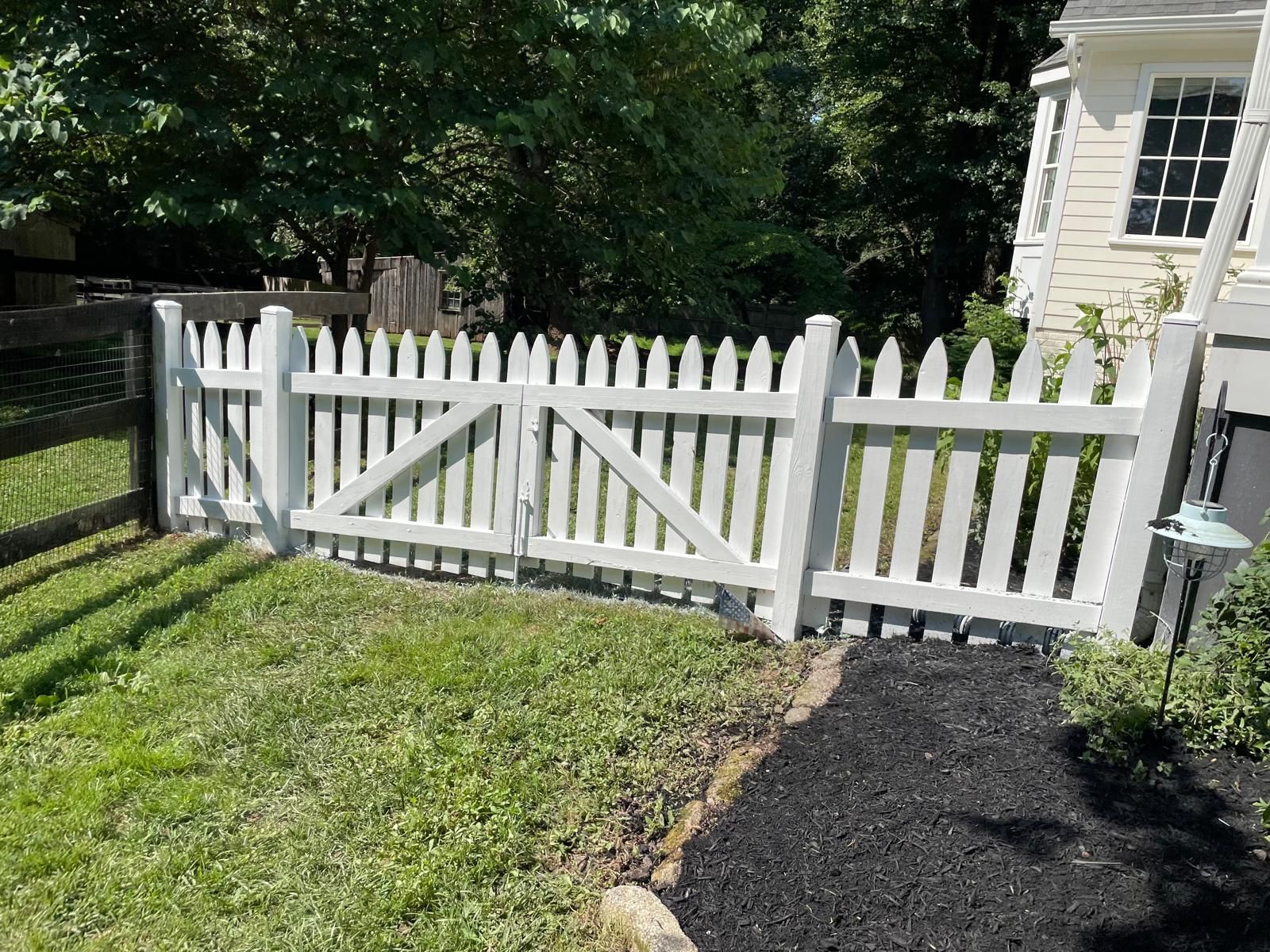 White picket fence with a gate beside a grassy yard and black mulch, in front of a house and trees.