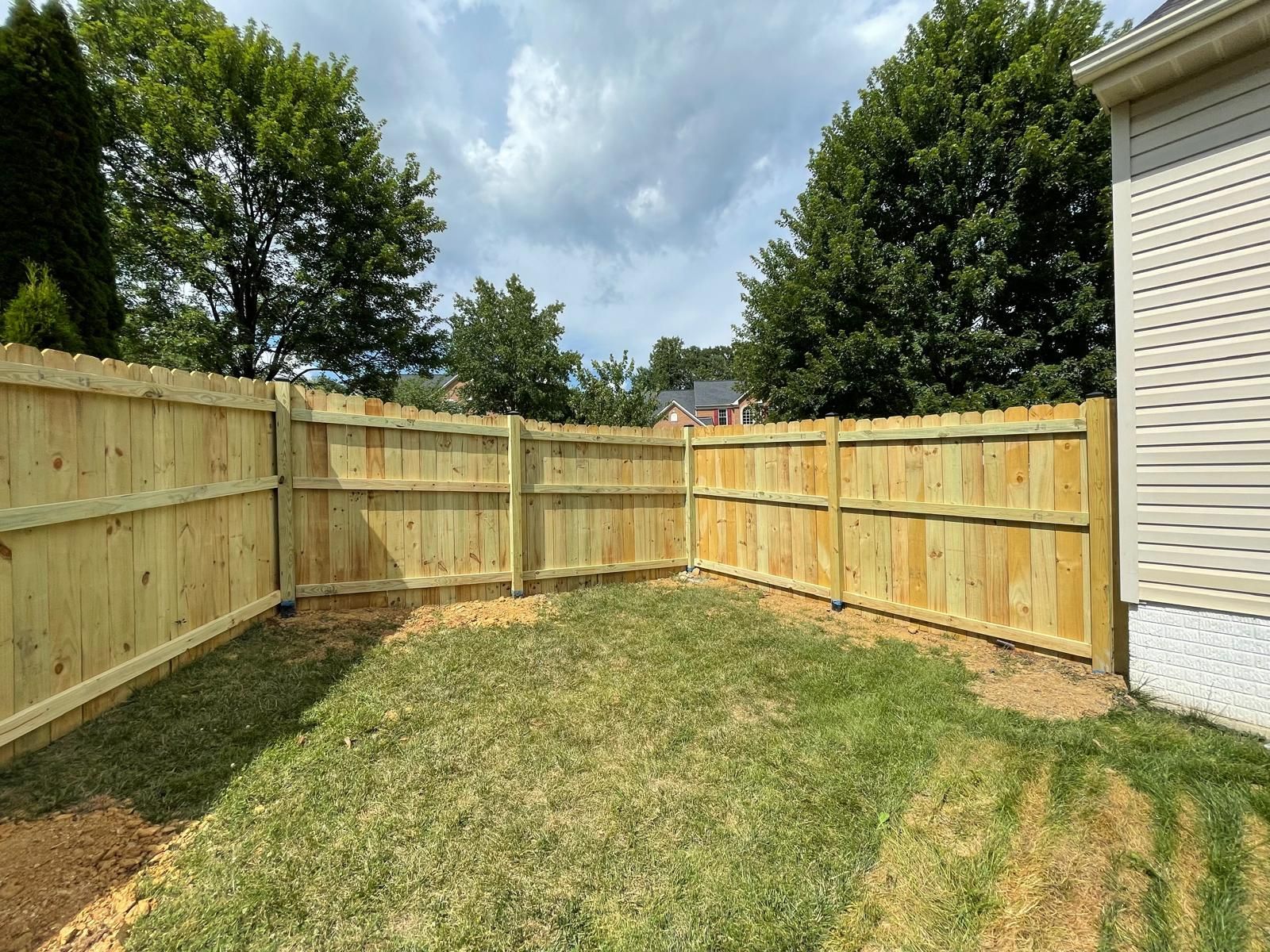 Backyard with new wooden privacy fence, patchy grass, and cloudy sky beside a house.